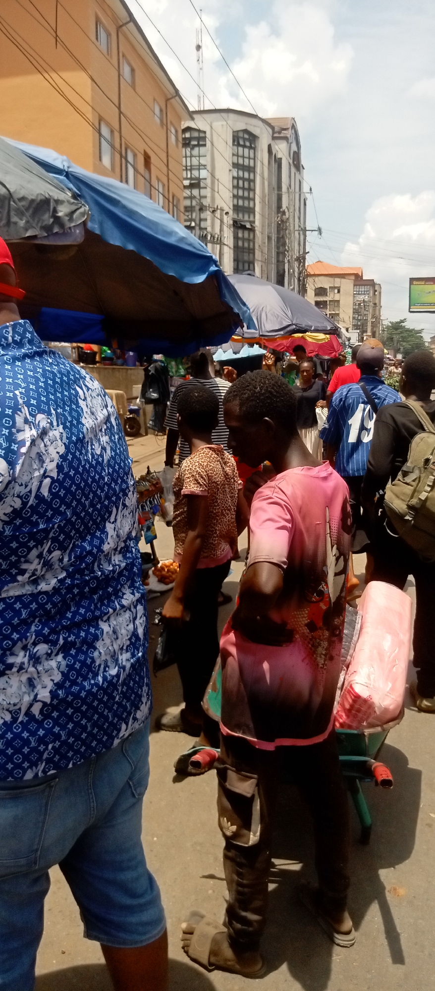 This media file captures a bustling outdoor market scene in broad daylight, likely on a street in Onitsha, Nigeria. The image is taken from a low angle, looking forward into a crowd of people.
In the immediate foreground on the left, the back of a person wearing a blue and white patterned short-sleeved shirt (resembling a Louis Vuitton monogram) and blue denim shorts is visible. To the right of them, a young person, possibly a vendor, is seen from the back and side. They are wearing a pink and black tie-dye t-shirt, dark trousers with an Adidas logo visible on a pocket, and sandals. This individual is pushing or standing next to a small green handcart containing a pink item wrapped in plastic.
Numerous other people populate the scene, walking and milling about. Many are casually dressed, with examples including a person in a leopard print top and another in a blue shirt with the number "19" printed on the back. In the mid-ground, a person can be seen near a small display of orange-colored goods, possibly fruits, on a white cloth.
The market area is shaded by several large, colorful market umbrellas (blue, grey, black), indicating vendor stalls. In the background, multi-story buildings with various architectural styles are visible, including one prominent light yellow/orange building and another taller, grey-white building with a complex window pattern. Overhead electrical wires crisscross the sky. The sky above is bright with scattered white clouds, suggesting a sunny day. Shadows on the ground confirm the sun is high. A green billboard or sign is partially visible high on the right side of the frame, but any text on it is unreadable. The overall atmosphere is one of a busy, active commercial environment.