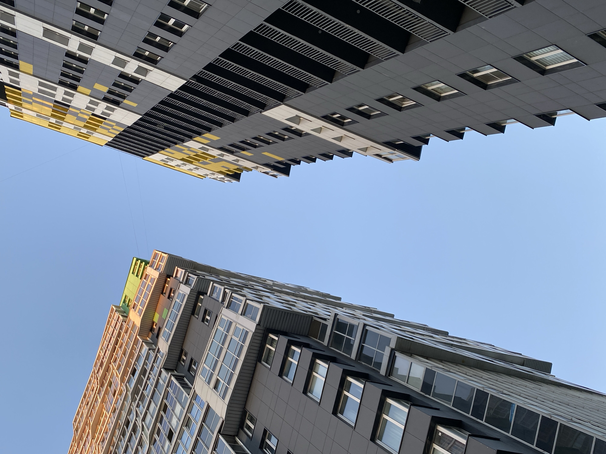 This is a low-angle, full shot of two modern apartment buildings under a clear blue sky. The buildings are tall and appear to be close to each other, with a view of the sky between them.
The building on the left is lighter in color, with a facade that appears to be a mix of grey and light beige panels, interspersed with numerous windows. The top section of this building features more colorful elements, including green and yellowish-orange sections, and its structure is more angular and stepped. Some balconies or recesses are visible.
The building on the right is predominantly dark grey, with accents of yellow and white in a geometric pattern on its upper sections. This building features many balconies with black railings. The windows are smaller and more uniformly placed compared to the building on the left.
The scene is set in the city of Kudrovo, Russia, a developing residential area known for its contemporary architecture. The image captures the architectural style of newer construction in the city. There are no people or animals visible in the image. The environment is urban, with a focus on the design and scale of the apartment buildings.