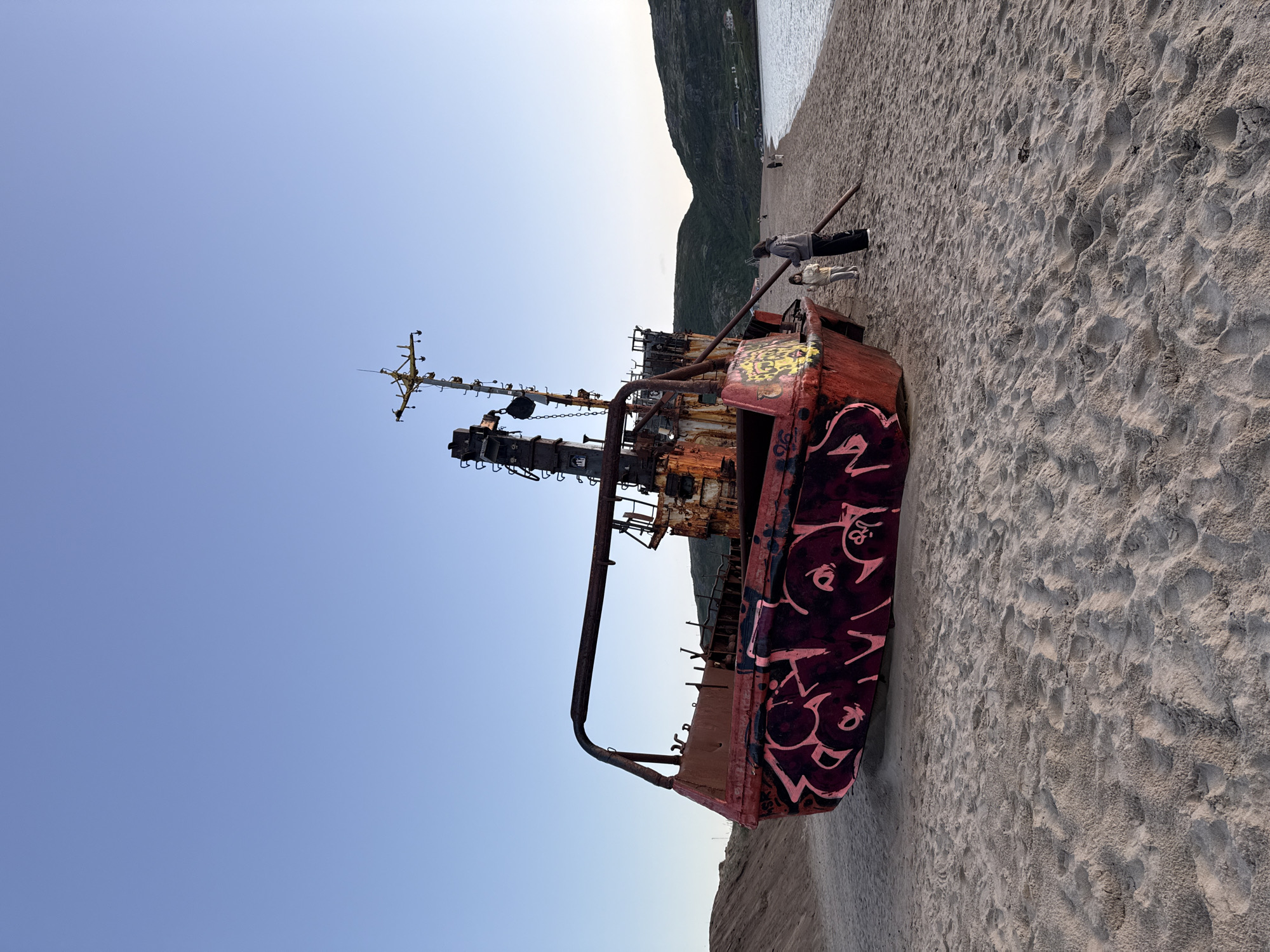 The image shows a rusty shipwreck with graffiti on the beach in Teriberka, Russia. The shipwreck is the prominent object in the foreground, tilted on its side, with its red hull covered in stylized pink and black graffiti. The ship has a tall, skeletal mast with various antennas and rigging. To the right of the shipwreck, two people, one adult and one child, are standing on the sandy beach, looking towards the ship or the sea. The beach stretches out behind them, meeting a body of water on the right side of the frame. In the background, rolling, green-covered hills rise up, with a few small structures visible on their slopes. The sky above is a clear, pale blue, suggesting it is either early morning or late evening. The sand is textured with footprints.