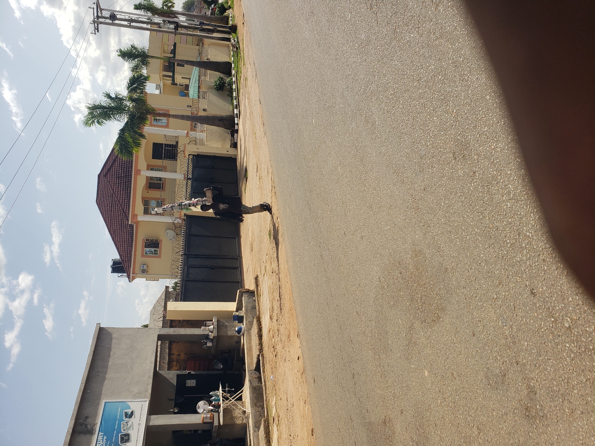 The image depicts a street scene in Gura Top, Nigeria, under a partly cloudy sky. In the foreground, a paved road takes up the majority of the frame, with a dirt shoulder on the right. On the left side of the road, a light gray building with a concrete facade houses a business called "Mercy Store," which advertises "Fashion Wear." Outside the store, a white folding table holds a fan and various items, and a small, light-colored building section with a dark doorway is adjacent. Further down the street, a large, two-story yellow house with a red-tiled roof stands behind a black metal gate topped with barbed wire. Several palm trees are planted in front of the house, and a utility pole with electrical wires is visible on the right. A man, dressed in a dark jacket and light-colored pants, is walking down the street carrying a long, narrow object balanced on his shoulder, appearing to be part of a ladder or scaffolding. He is walking away from the camera and towards the residential building.