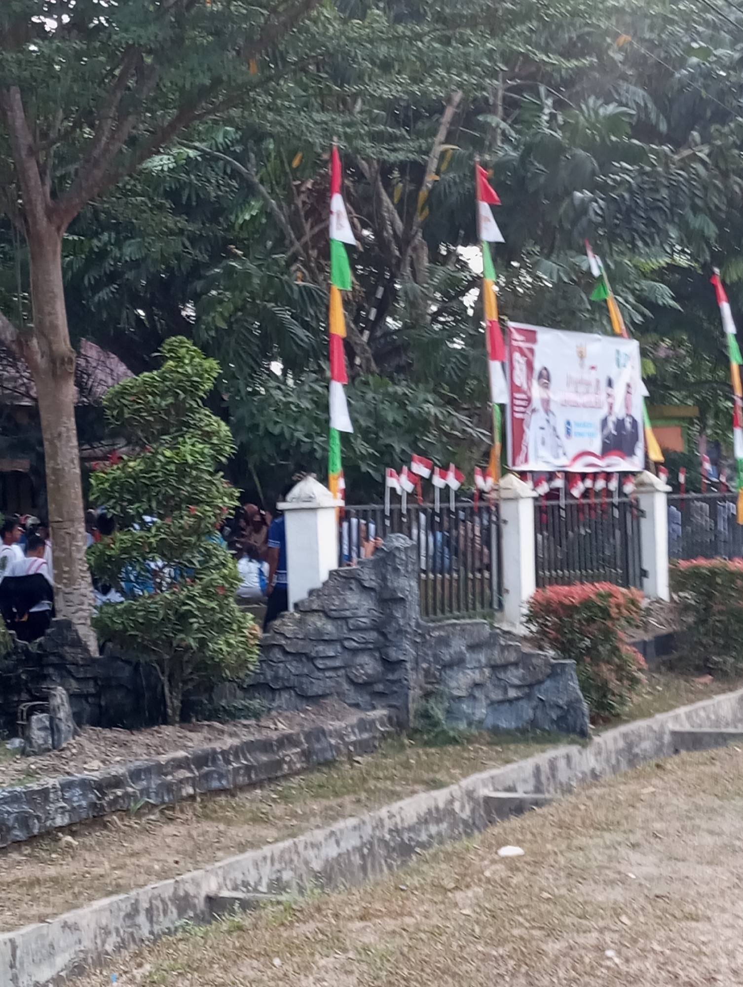 The image shows an outdoor scene in Pematang Reba, Indonesia, likely in front of a building or facility decorated for a public event. Multiple Indonesian flags, red and white, are displayed on poles along a fence. A large banner with images of men in formal attire and text in Indonesian is prominently featured. The banner appears to be a promotional or informational display. In the foreground, there are stone-like structures and landscaping with green foliage and small red-leafed bushes. People are visible in the background, some appearing to be attendees or participants in the event, and others are walking. The environment suggests a public gathering or celebration.