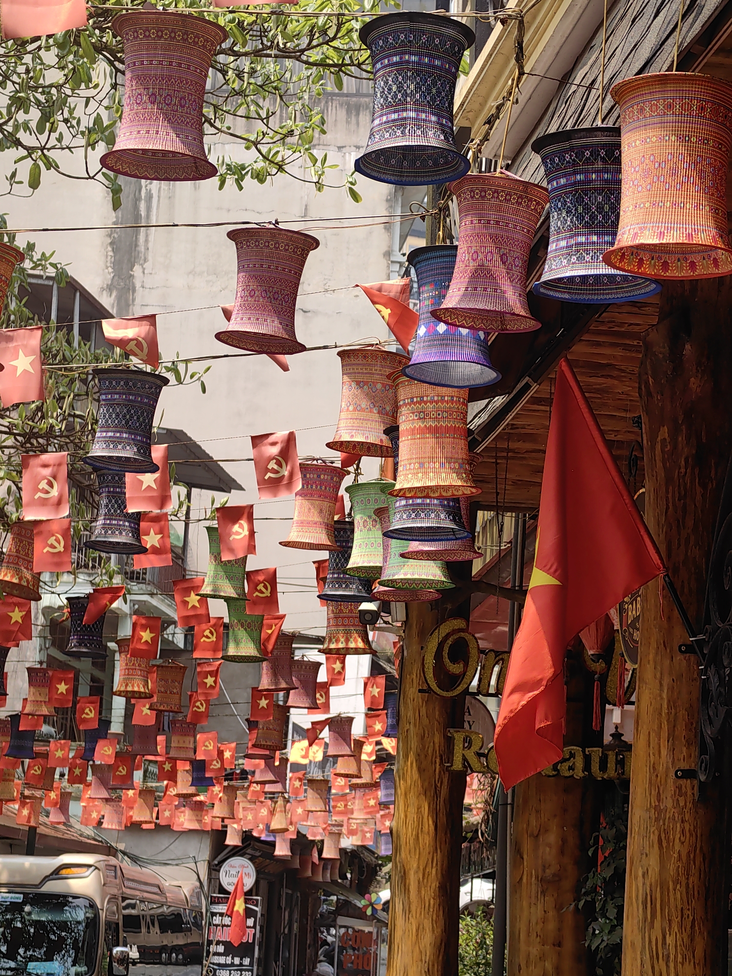 This image captures an outdoor street scene in Sa Pa, Vietnam, characterized by extensive overhead decorations. Numerous barrel-shaped, intricately patterned woven objects, resembling lanterns or shades, are suspended by wires across the thoroughfare. These decorations display a variety of colors including red, purple, blue, green, and orange, featuring geometric and floral patterns.
Interspersed among these woven objects are dozens of small red flags. Some flags display a yellow five-pointed star, representing the Flag of Vietnam, while others feature a yellow hammer and sickle, indicative of the Communist Party of Vietnam flag. A larger, unfurled red flag with a yellow star is affixed to a vertical wooden pillar on the right side of the frame. This pillar is part of a building with prominent wooden architectural elements; partial signage for "Oma" and "Restaurant" is visible on this structure.
On the left side of the street, the front of a light-colored bus or van is partially visible, reflecting the surroundings. Further down the street, additional commercial signage, including "Nail Mi" and "Hair Cut," can be discerned. Green foliage from tree branches overhangs the street, intertwined with the suspension wires for the decorations. The background features multi-story buildings with light-colored facades. The scene is illuminated by bright daylight.