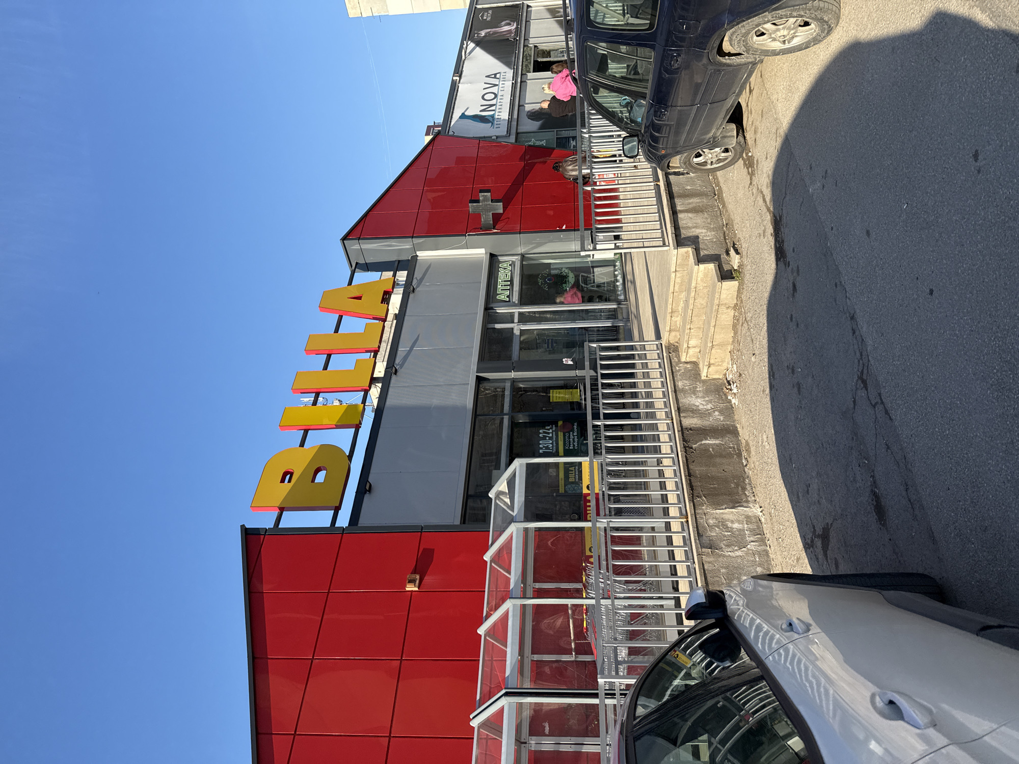 The image shows the front of a Billa supermarket building in Sofia, Bulgaria. The building has a red and gray exterior with the "BILLA" logo prominently displayed in yellow and red lettering. An "apteka" (pharmacy) sign is visible near the entrance. There are two vehicles parked along the side of the building - one silver and one navy blue. Shoppers are visible inside the building and on the second level. The sky is clear blue.