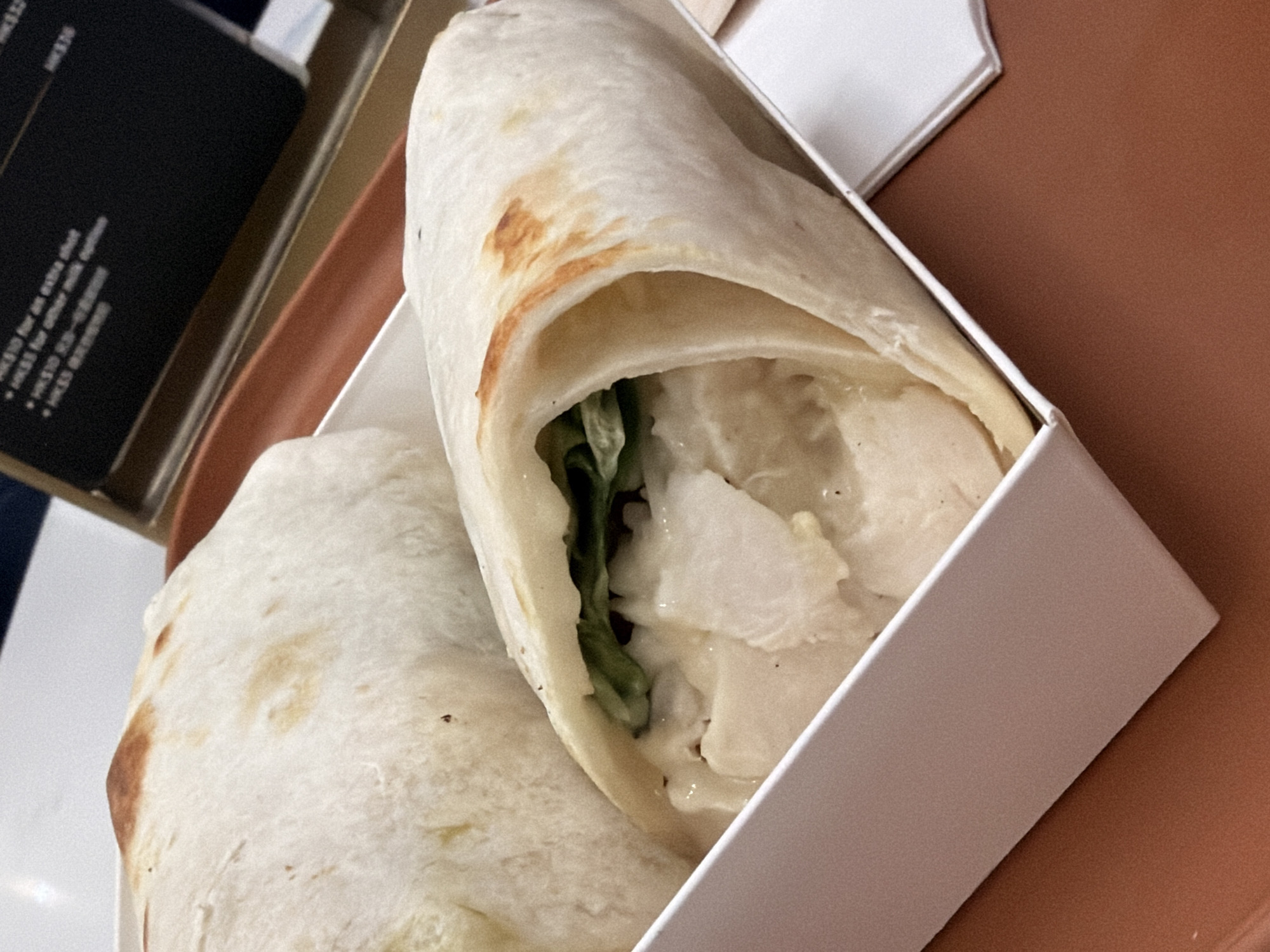 The image shows two chicken wraps on a brown tray, likely in an airplane cabin as indicated by the text in the background which appears to be a menu with prices in Hong Kong dollars (HKD) and the phrase "extra shot." The wraps are made of white tortillas and are filled with chicken, lettuce, and a creamy sauce. The wraps are presented in a white rectangular container. The lighting is artificial and appears to be from above. There are no people visible in the image. The overall scene is one of in-flight catering.