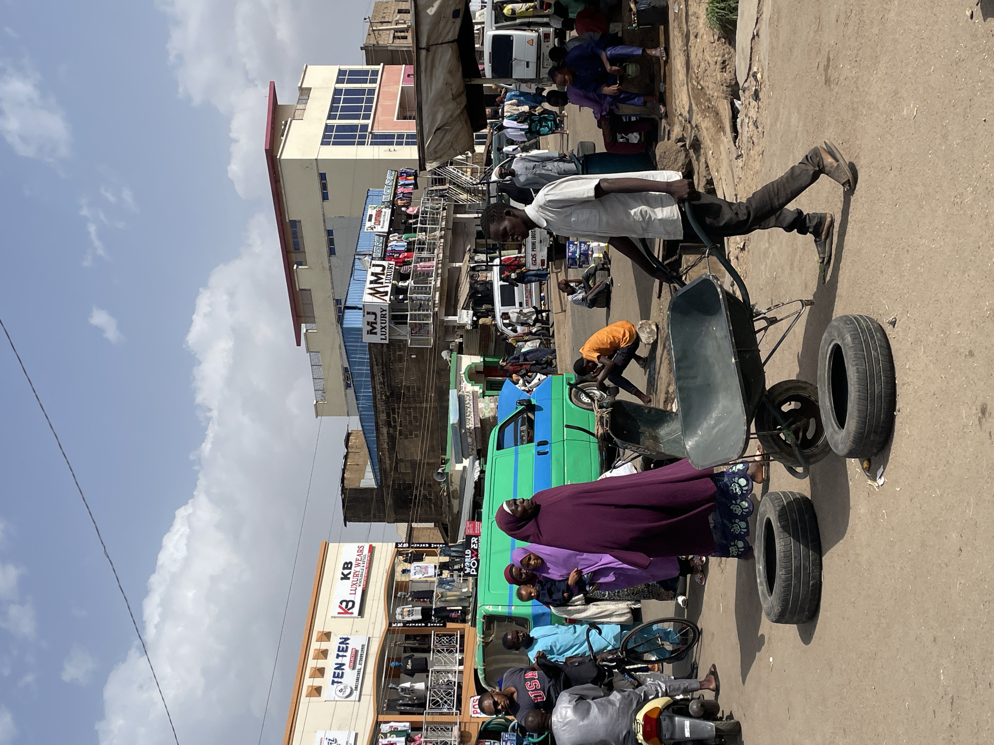 This image captures a vibrant and busy street scene in Jos, Nigeria, during daylight hours under a sky with scattered white clouds. The setting is a commercial area with multi-story buildings lining the street, housing various businesses.
Numerous people are visible, engaged in daily activities. In the mid-ground, a man in a white shirt and dark trousers pushes a unique green wheelbarrow or cart equipped with two large vehicle tires attached to its rear frame. Near him, several individuals, including women wearing hijabs and long dresses (one prominently in purple), stand beside a distinctive green and blue utility vehicle or bus. A child in an orange shirt is bent over near the ground. In the foreground, a person sits on a motorcycle. The street itself appears to be a mix of dirt and paved sections.
The commercial nature of the area is evident from the numerous shop signs. Visible text includes "TEN TEN COLLECTION" with phone numbers "08025803953, 07069919905," "KB LUXURY WEARS WHOLESALE & RETAIL," "MJ LUXURY," and "WAZIRI SPORT." Other signs read "WORLD POWER," "GDS PRIME LOGIS," and "WOWN AUTOSPEED." A man is seen wearing a shirt with "USA" printed on it. The scene depicts a typical bustling urban environment with a mix of pedestrian traffic, handcarts, motorcycles, and other vehicles.