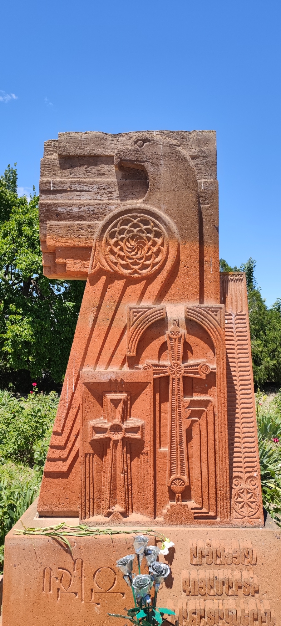 This is a full, eye-level, vertical, outdoor shot of a large, reddish-brown stone monument in Araks, Armenia. The monument is intricately carved with various symbols, including what appears to be a bird at the top, a circular floral design, and multiple crosses. The stone has a rough texture and shows signs of weathering. At the base of the monument, there are inscriptions in Armenian and a small bouquet of silver-colored artificial roses with a few white artificial flowers. The monument is set against a backdrop of lush green trees and a clear blue sky. The lighting suggests it is daytime, with shadows cast by the carvings indicating the sun is to the left.