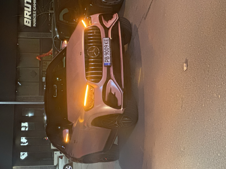 A light-colored Mercedes-Benz GLE Coupe SUV is parked at night, viewed from the front, angled slightly toward the right. The vehicle's LED daytime running lights/turn signals and side mirror indicators are illuminated with an orange glow. Its front grille features prominent vertical slats and a large Mercedes-Benz emblem. The Bulgarian license plate "PB 9009 KE" is affixed to the front bumper. The vehicle is situated on a dark paved surface, where a small, light-colored rectangular object lies on the ground in the lower right foreground. In the background, a commercial building is visible with large windows. A partially illuminated sign on the building to the right reads "BRUT..." and "NARGILĖ GAMING" below, identifying the establishment as "Brutal Nargile Gaming." A tall, red, stylized giraffe sculpture is positioned behind the Mercedes and to the right, visible against the building. Other vehicles are partially visible, including a light-colored sedan to the far left and a dark-colored vehicle to the right of the Mercedes. No people are present in the scene. The license plate prefix "PB" and the specific business signage confirm the location as Plovdiv, Bulgaria.