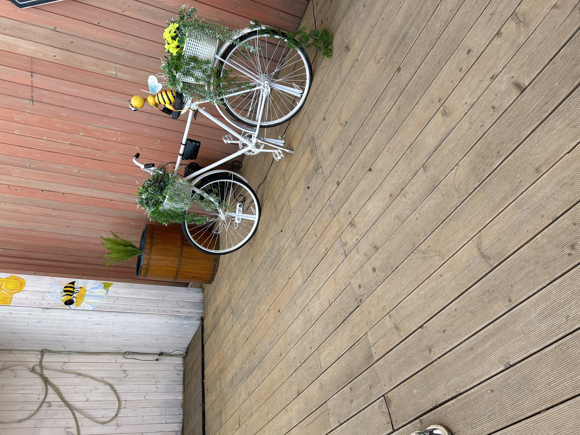 The image shows a decorated white bicycle with baskets holding flowers. It is positioned on a wooden deck near a wall decorated with a painted bee and a wooden barrel containing a fern. The location is Chaikovsky, Russia.