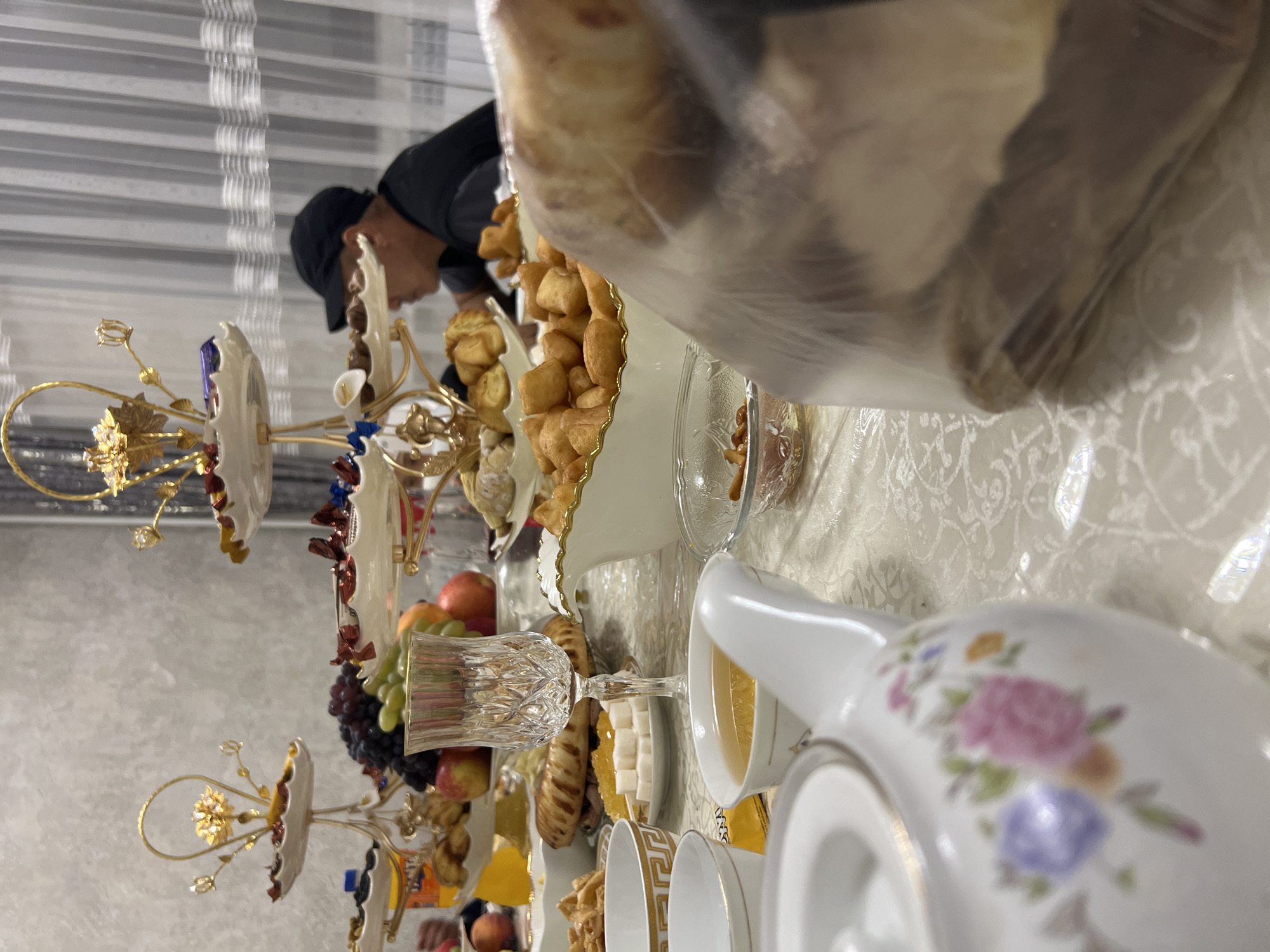 The image depicts a table laden with food and drinks, suggesting a gathering or a meal. In the foreground, a white teapot with floral decorations is prominent. Beside it, a small white cup contains a golden-colored liquid, likely tea. Further back, a clear glass goblet sits on the table, and a plate of white sugar cubes and some orange candies is visible. Several tiers of serving dishes are arranged on the table, holding various food items including fruits like grapes and apples, and what appear to be pastries or fried dough. In the background, a person wearing a black baseball cap and dark clothing is partially visible, facing away from the camera. The setting appears to be indoors, with a curtained window in the background. The overall impression is that of a festive or celebratory meal. The location is identified as Төмөнкү Ала-Арча, Kyrgyzstan.