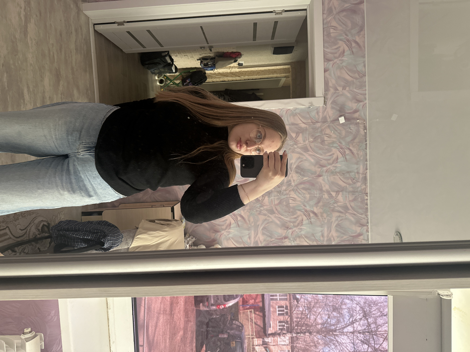 A young woman is captured in a mirror selfie indoors, posing slightly angled to her left. She has long, light brown hair, wears round glasses, a black long-sleeved top, and light blue jeans. She holds a dark smartphone, likely an iPhone, in her right hand to take the photo.
The reflection in the mirror reveals a room or hallway with light-colored walls and a closed white door featuring two horizontal panels. Various items are scattered on the floor or a low surface in the background, including bags, a green folding chair or stool, and other miscellaneous objects. The wall immediately to the right of the mirror has a distinctive pink and grey patterned wallpaper.
Through a window in the foreground, an outdoor urban scene is visible, consistent with Chaikovsky, Russia. Bare tree branches suggest either late autumn, winter, or early spring. Several multi-story buildings with numerous windows, a street or parking area, and a vehicle are discernible. A white radiator is partially seen below the window. The lighting appears to be natural daylight, possibly under overcast conditions. No visible text is present in the image.
