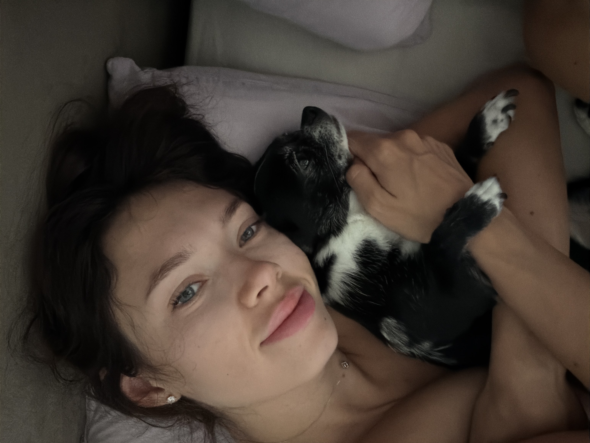 A person with dark hair and blue eyes, wearing a delicate necklace, is lying in bed and embracing a small black and white dog. The person's head is resting on a pillow. The dog is looking to the right. The scene appears to be indoors, possibly in a bedroom. There is no specific indication of the location within Tel-Aviv, Israel.