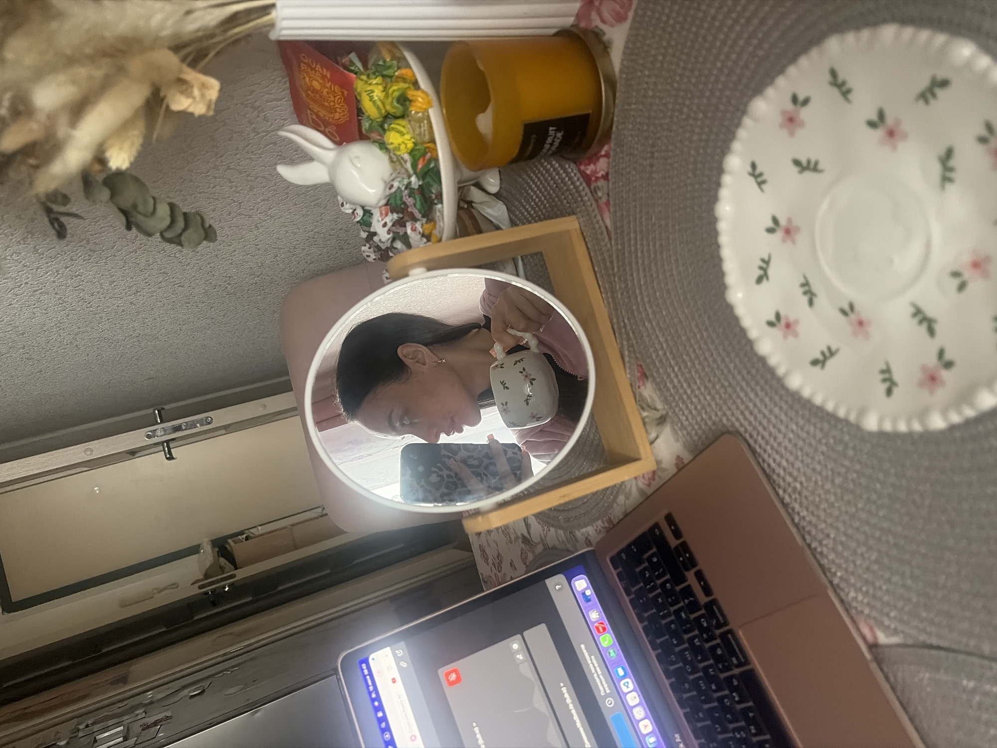 The image depicts an indoor scene, likely a home office or living area, with a focus on a woman and her immediate surroundings. The woman is seen in profile, reflected in a circular mirror, holding a small, floral-patterned teacup to her lips. She is wearing a light pink jacket. The mirror itself is propped on a wooden stand. To the left of the mirror, a rose-gold laptop is open, its screen displaying what appears to be a website or application. To the right of the mirror, on a patterned surface, are several decorative items including a white ceramic bunny figurine, a small white vase with dried floral arrangements, a stack of colorful wrapped candies, a red packet with Chinese characters, and a lit orange candle in a glass jar. In the foreground, a grey placemat is visible, underneath which is a white saucer with a similar floral pattern to the teacup. The background shows a doorway and a wall. The setting is illuminated by artificial light. The presence of the laptop and the woman's attire suggest a casual, possibly work-from-home or leisure activity. The overall impression is one of a cozy and personalized space.