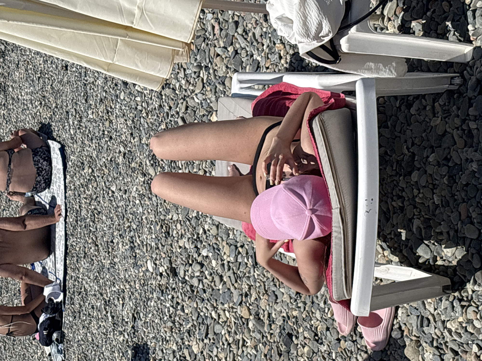 On a sunny day at a pebble beach in Sochi, Russia, a woman lies on a white lounge chair, her legs extended upwards. She is wearing a black bikini and a pink baseball cap that covers her face. A pink towel is draped over the lounge chair. To the left of the chair are two pink slides. In the background, other people are sitting on the beach. A portion of a cream-colored beach umbrella is visible on the right side of the image. The ground is covered in small grey pebbles.