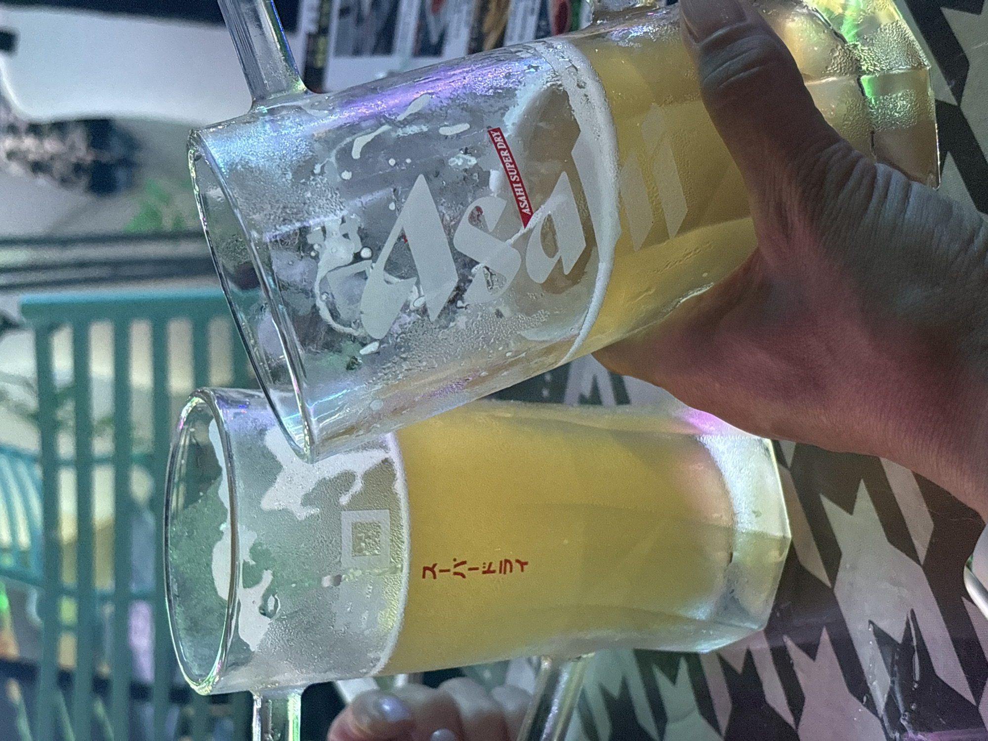 In a dimly lit establishment, two glasses of beer are being held up, one by a hand, in a toasting gesture. The beer is a pale yellow color and has a frothy head. The glasses are made of clear glass and are sweating from the cold. On the left glass, Japanese characters are visible, reading "Super Dry". On the right glass, a red and white label says "ASAHI SUPER DRY". The background is blurred but appears to be a seating area with a patterned surface on the table. The overall atmosphere suggests a casual social gathering.