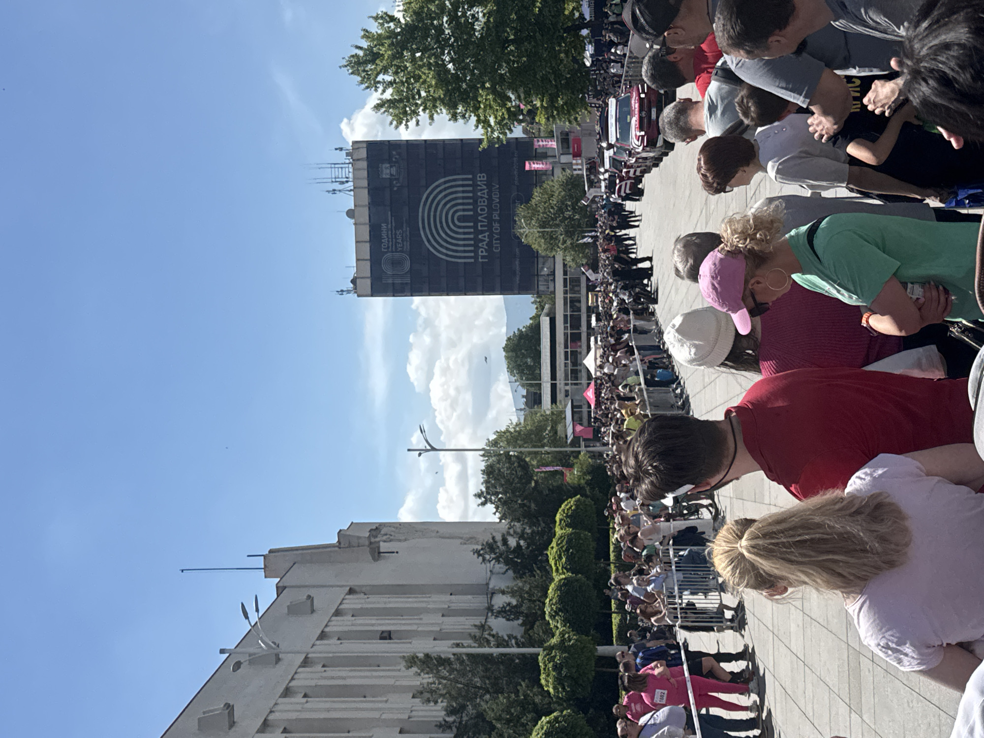 A crowd of people stand behind a metal barricade on a paved area. In the background is a building with a large banner reading "ГРАД ПЛОВДИВ CITY OF PLOVDIV", likely in Plovdiv, Bulgaria. There are trees and other buildings visible behind the crowd. Some individuals are wearing pink attire.