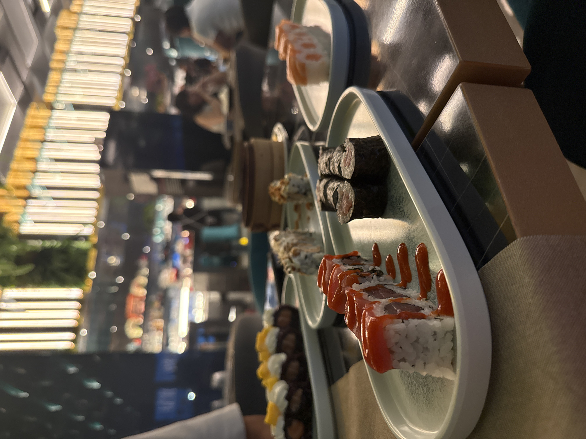 The photograph captures a vibrant scene inside a modern sushi restaurant, likely during evening hours, illuminated by artificial light. The image is taken from a diner's perspective, with various sushi plates visible on a conveyor belt system.
In the foreground, several distinct plates of sushi are sharply in focus. The closest plate on the right displays sushi rolls with white rice on the outside, topped with slices of red fish (likely tuna or salmon) and drizzled with a reddish, spicy-looking sauce. To its left, another plate features dark, nori-wrapped maki rolls. Further along the conveyor, additional plates showcase other varieties of sushi, including rolls with lighter toppings and some with yellow and white garnishes. A bamboo steamer basket, possibly containing dumplings, is also faintly visible in the midground.
The light-colored plates, possibly white or light gray, rest on a brown, multi-tiered structure, which forms part of the automated serving system. This structure has a slightly reflective surface.
The background is intentionally blurred, highlighting the passing food. Despite the blur, numerous bright, linear lights, resembling overhead or decorative LED strips, are discernible, casting a warm, yellowish glow. Silhouettes and indistinct forms of other diners and restaurant staff can be seen in the distant background, indicating a moderately busy environment. The overall ambiance suggests a casual yet refined dining experience centered around Japanese cuisine. No discernible text is visible within the image.
