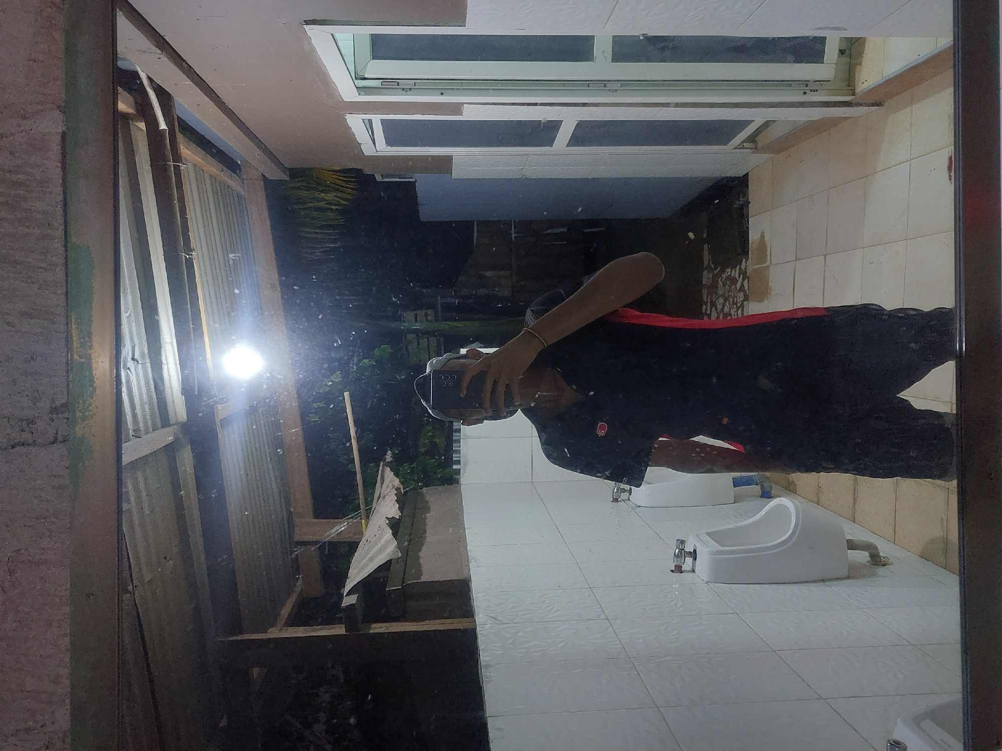 A mirror reflects a person in a black jacket with red trim, holding a cell phone in a tiled bathroom. The bathroom features multiple white urinals. Outside, a corrugated metal structure is visible. The location is Dar es-Salaam, Tanzania.