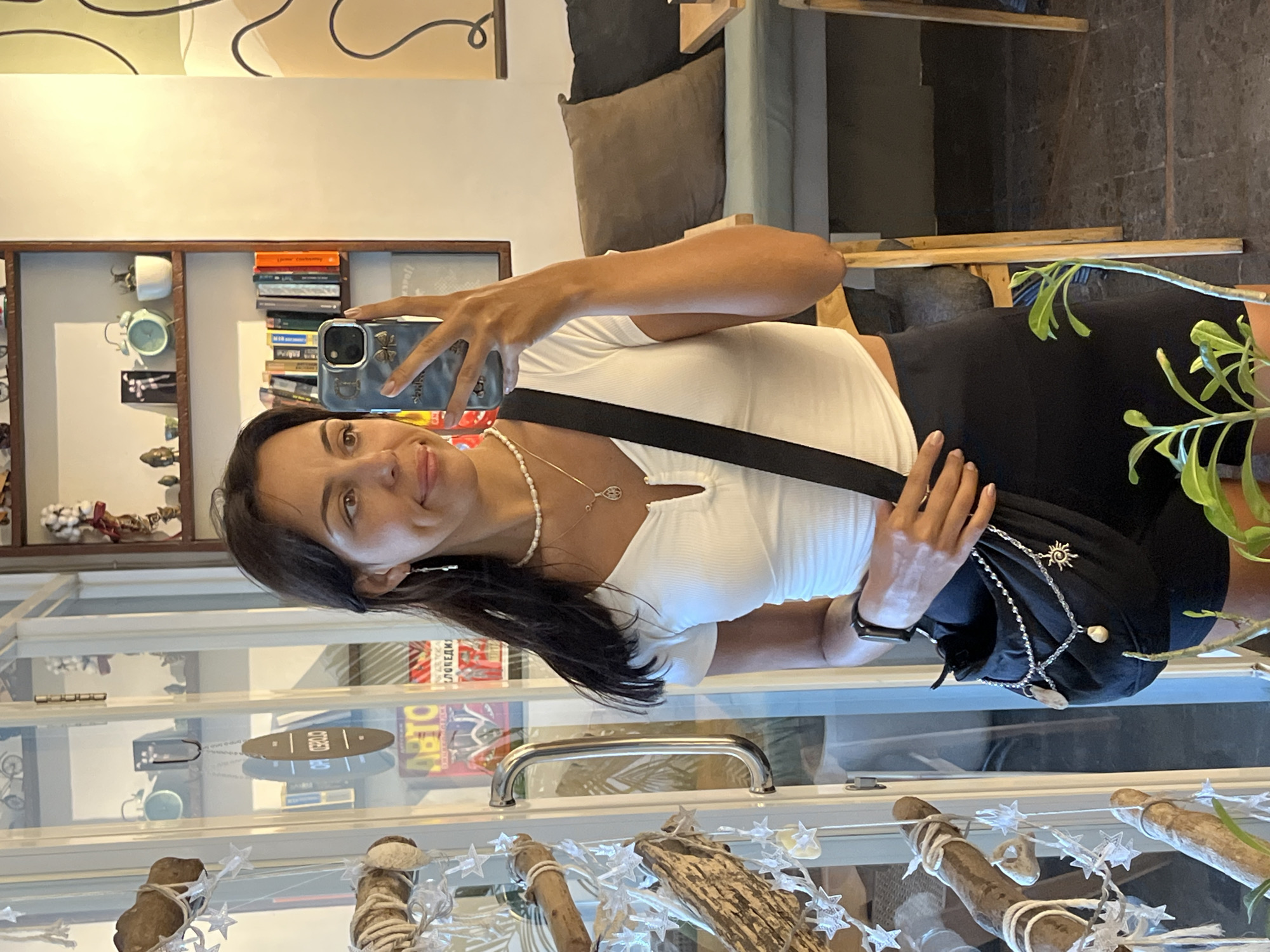 A woman takes a selfie in a Kuta Selatan, Indonesia, shop. The woman wears a white, v-neck shirt and black shorts with a black cross-body purse. The shop contains glass shelves with items displayed for sale.