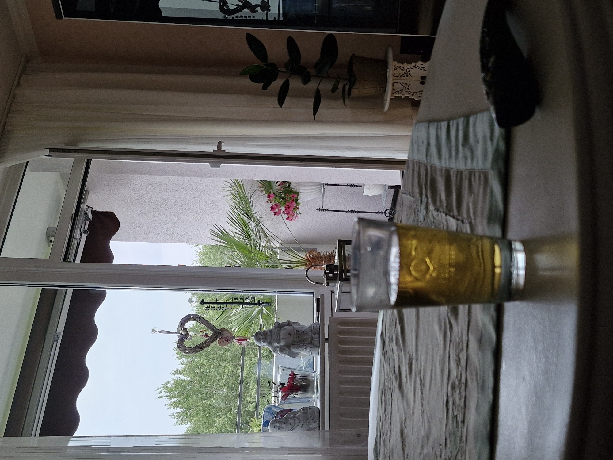 The image depicts an interior scene with a partial view of an outdoor balcony. In the foreground, a glass of golden liquid, likely beer, sits on a table. A dark object, possibly a remote control, is also on the table. Behind the glass, a textured runner is laid across a bed or sofa. Through a glass sliding door, a balcony is visible with a heart-shaped decoration hanging, trees in the background, potted plants, and two Buddha statues on a windowsill. A thermometer is also visible on the window frame. The room appears to be in a residential setting, likely an apartment. The ambient lighting suggests it is daytime. There are no people visible in the image. The specific location in Bremen, Germany is not determinable from the visual information provided.