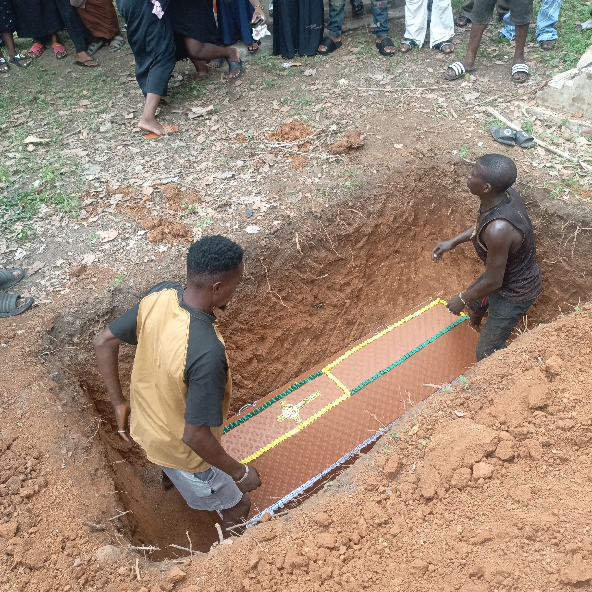 The image depicts two men inside a rectangular excavated pit in Andaha, Nigeria, lowering a brown and patterned casket. A group of people stand around the perimeter of the pit. The ground surrounding the pit is red soil mixed with dried leaves. The men are wearing casual clothes, and some people around the pit are wearing sandals or open-toed shoes.