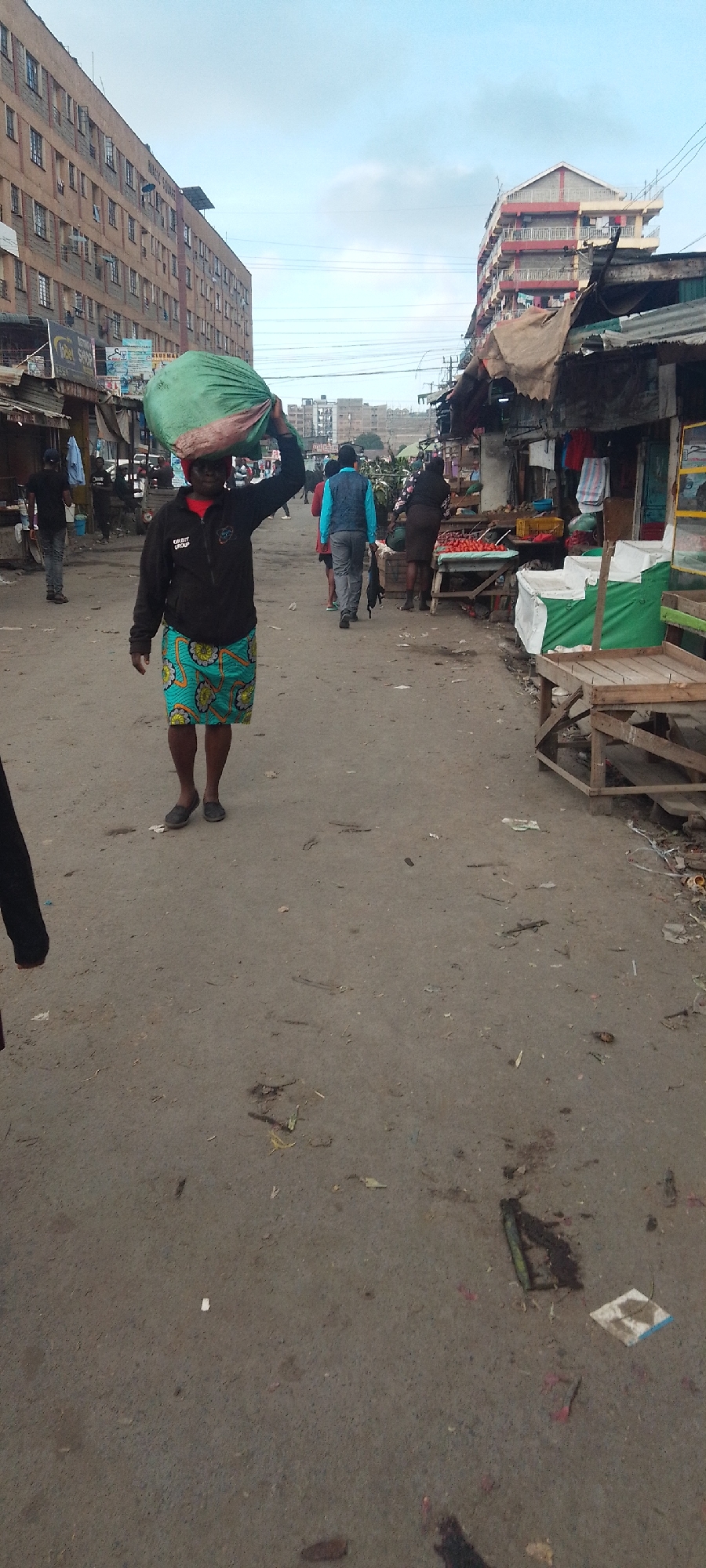 The image depicts a street scene in Nairobi, Kenya. An African woman in a black jacket, red shirt, and a blue and yellow patterned skirt carries a large green bag on her head. She is walking down a dirt road that appears to be a marketplace. There are several people in the background walking in the same direction, including a man in a blue vest. Stalls line the right side of the road, displaying produce and other items. The left side of the road has buildings with storefronts. The ground is littered with debris. Buildings in the distance show the urban landscape of Nairobi. The sky is overcast.