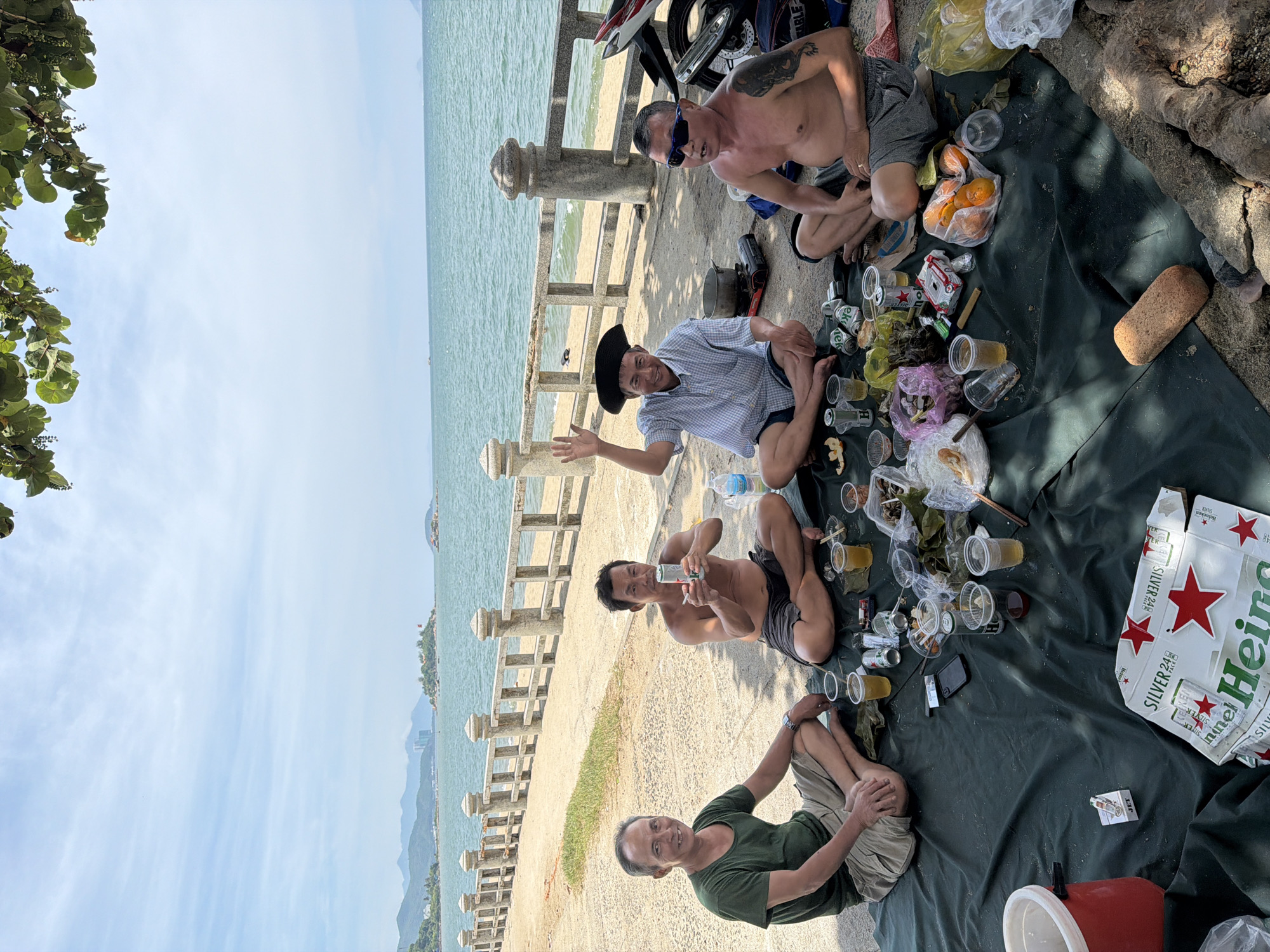 Four adult males are having a picnic on a dark green tarp on a sandy area next to a paved walkway. The walkway has a stone railing and a view of the sea and distant islands. The men are sitting in a circle, with food and drinks spread out between them. The man on the far left is wearing a green t-shirt and beige shorts. The man to his right is shirtless, wearing dark shorts, and holding a can. The third man from the left is wearing a blue and white checkered shirt and a black hat, and is waving. The man on the far right is shirtless, wearing grey shorts, and sunglasses, and has a tattoo on his left arm. There are several plastic cups with beverages, food items in plastic bags, an open beer box, a red cooler, a portable stove, and a motorcycle in the background. The scene appears to be in Nha Trang Ward, Vietnam, given the coastal environment and likely demographics.