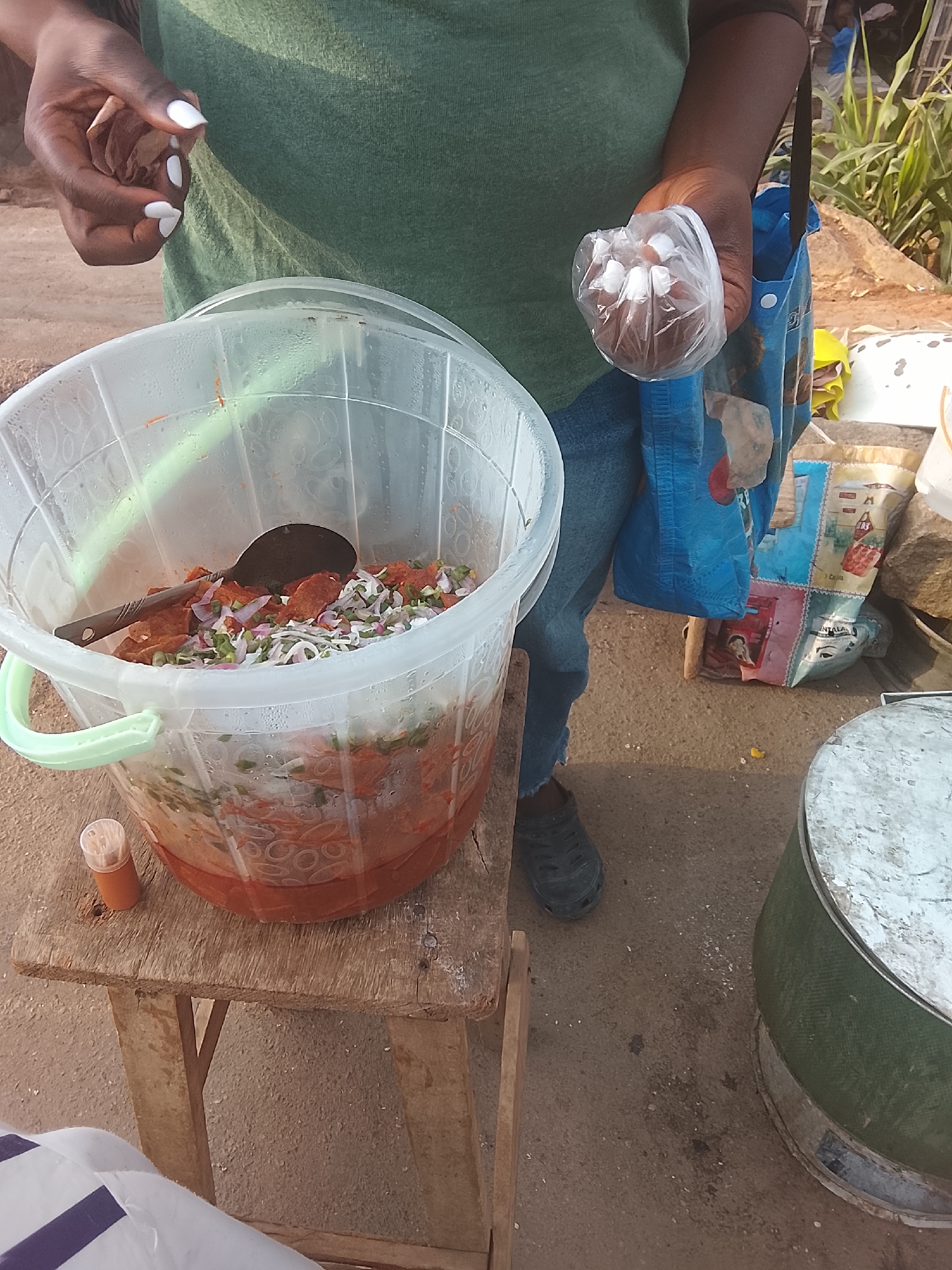 A street vendor in Gwong, Nigeria, stands next to a plastic bucket containing a layered mixture of ingredients, including a red sauce, white vegetables, and cut pieces of what appears to be meat. The bucket sits atop a small wooden stool. A small vial of orange liquid stands next to the bucket. The vendor holds a small plastic bag in one hand. The vendor wears a green shirt and blue jeans. A blue shopping bag hangs off the vendor's shoulder. A green drum with a white top sits next to the vendor's foot. The ground is dirt.