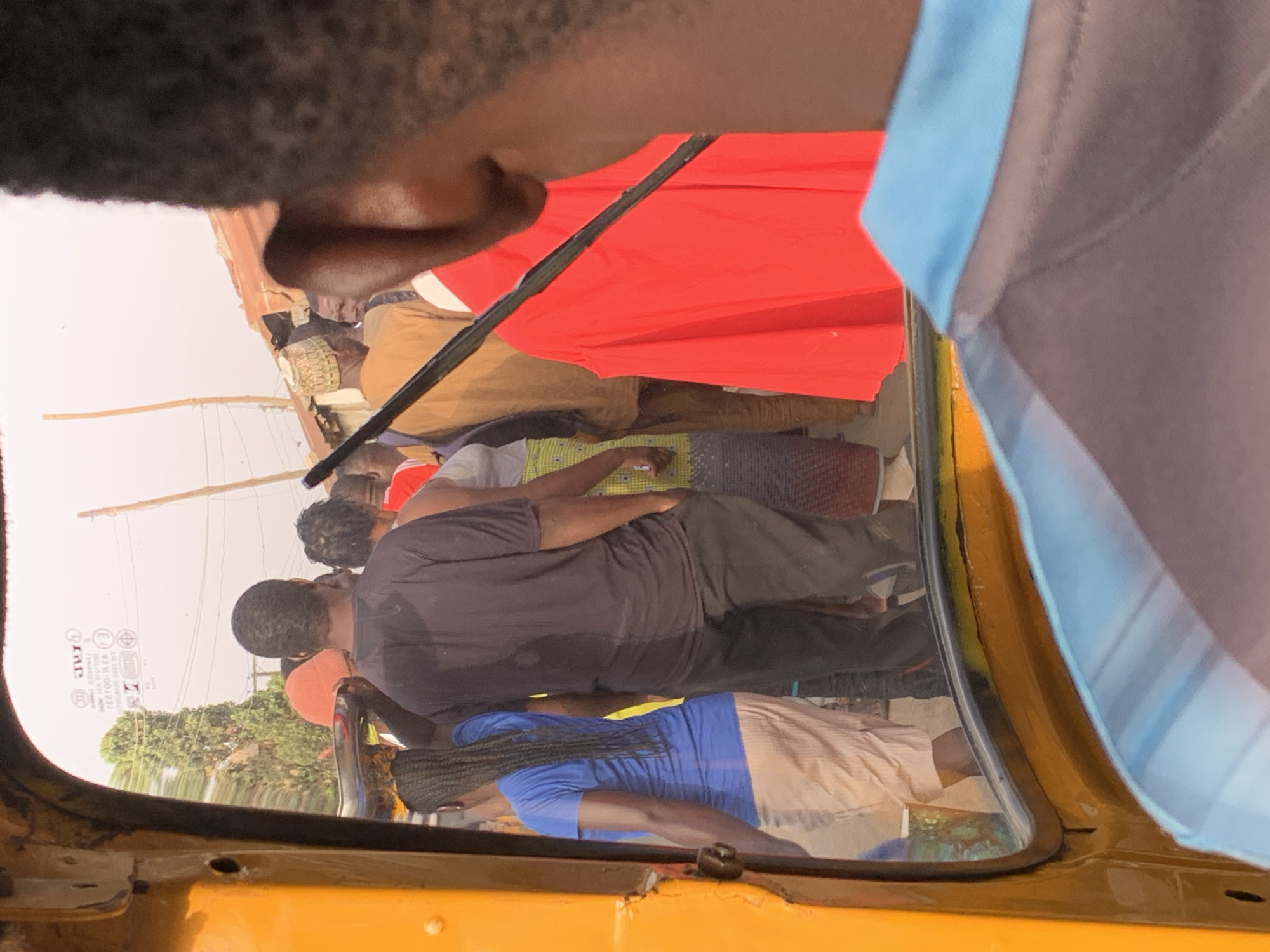 From inside a yellow vehicle, possibly a tuk-tuk, a street scene is visible through the windshield. A group of people are gathered outside, including individuals wearing a red garment, a black shirt, and a blue shirt. The environment includes trees and power lines. The location is inferred to be Jos, Nigeria.