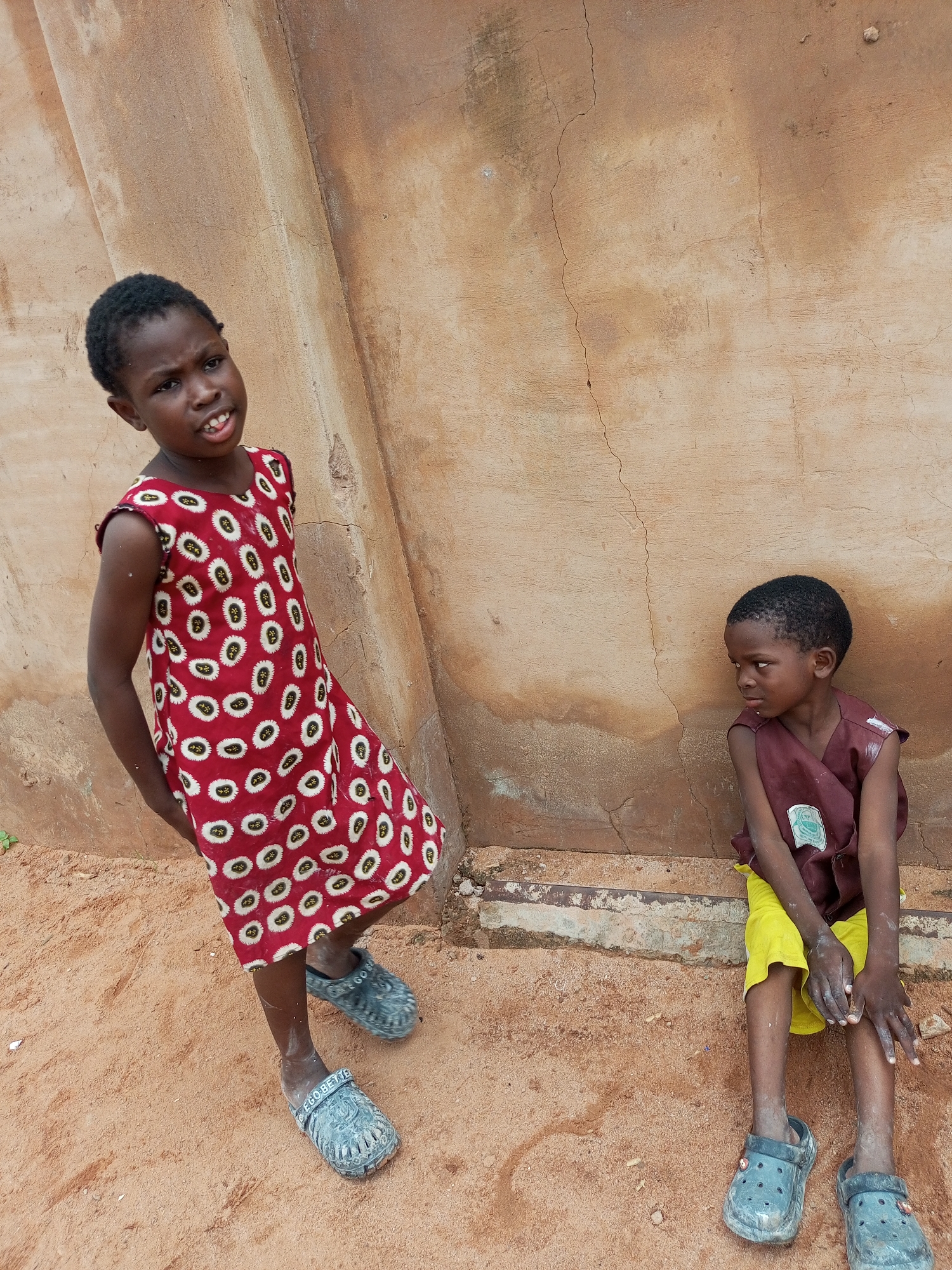 Two young children are positioned near a textured, weathered, beige wall in what appears to be an outdoor urban setting in Tempure, Nigeria. The child on the left, a girl, stands facing forward and slightly to the left, wearing a red sleeveless dress with a pattern of white oval shapes containing yellow centers. Her hands are behind her back. She has dark hair and is looking directly at the camera with a curious expression. She is wearing grey clogs. The child on the right, a boy, is seated on a low ledge, facing to the left and looking away from the camera. He is wearing a dark maroon sleeveless garment over bright yellow shorts. A patch is visible on the maroon garment. He is also wearing grey clogs. The ground beneath them is covered in reddish-brown sand or dirt. The wall behind them has visible cracks and stains, suggesting age and exposure to the elements. The overall environment appears to be simple and possibly impoverished.