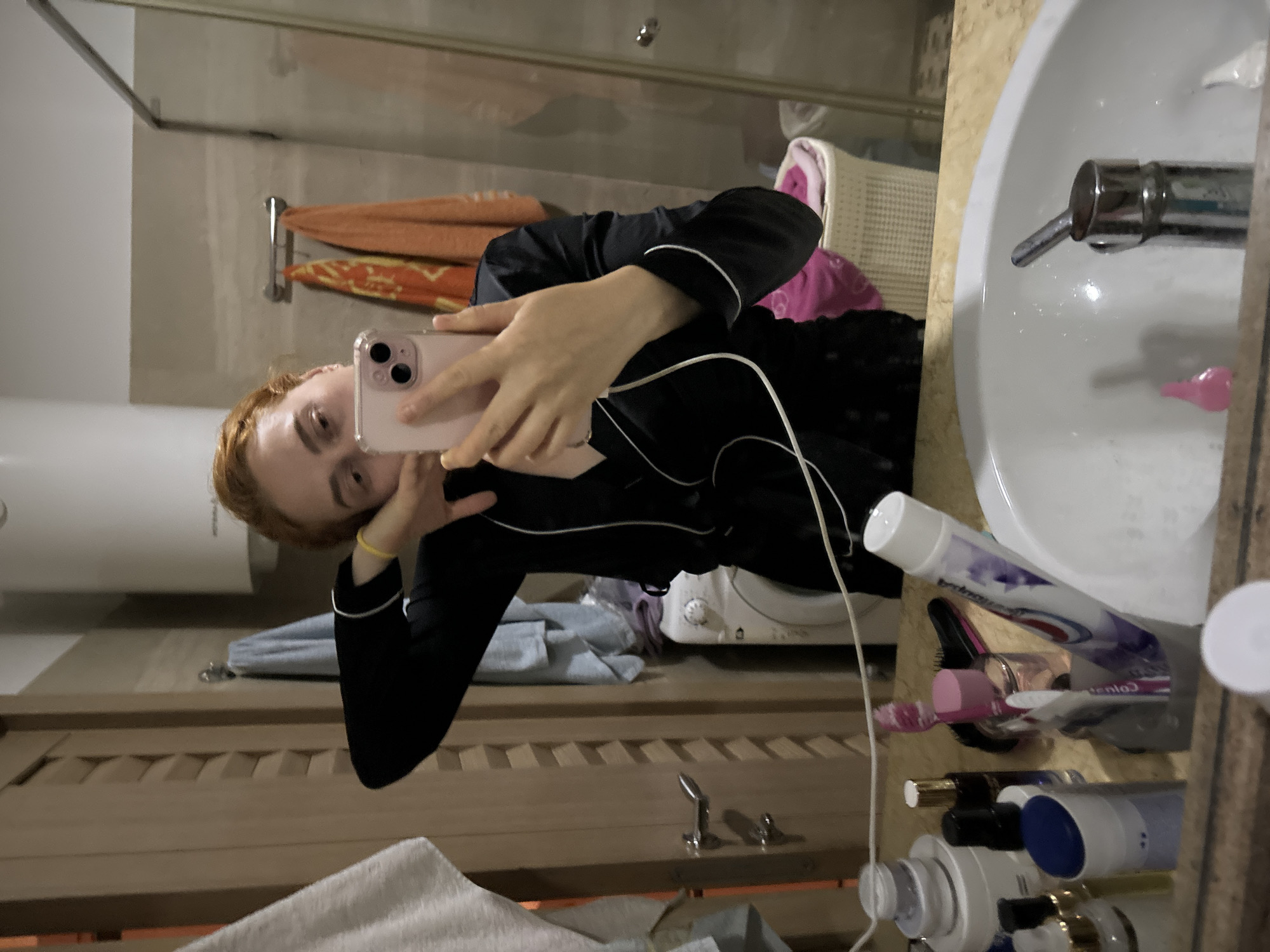 A young woman with red hair is taking a selfie in a bathroom. She is wearing black pajamas with white trim. The bathroom has a sink, a mirror, a washing machine, a towel rack with towels, and various toiletries. The location is Budva, Montenegro.