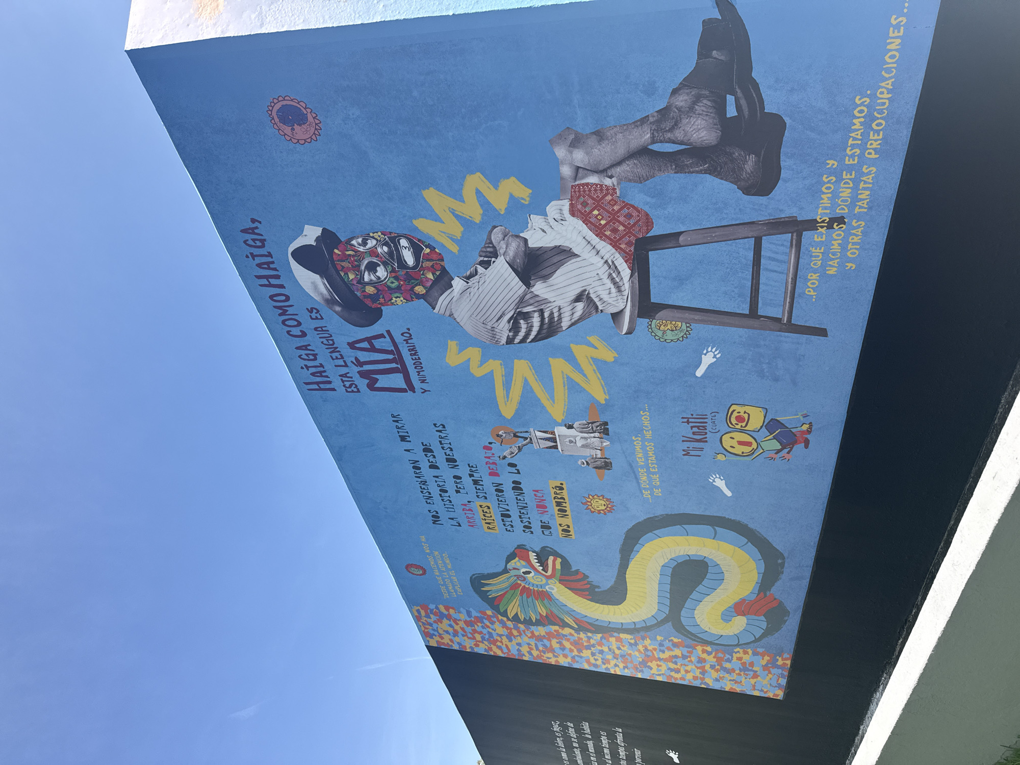 The image captures a vibrant mural located in San Andrés Cholula, Mexico. The mural is painted on a wall and features a variety of artistic elements. At the top, there is a collage-like image of a figure with a patterned mask, legs perched on a stool, and zig-zagged lines around it. Below, a colorful serpent or dragon-like figure stretches across the lower portion of the mural. There are also hand-drawn-style illustrations, text, and symbols scattered throughout the composition. The text is in Spanish and includes phrases such as "HAIGA COMO HAIGA," "...DE DÓNDE VENIMOS. DE QUÉ ESTAMOS HECHOS...," and "...POR QUÉ EXISTIMOS Y NACIMOS, DÓNDE ESTAMOS. Y OTRAS TANTAS PREOCUPACIONES..." The mural reflects a cultural and artistic expression, possibly exploring themes of identity, history, and existential questions. The sky is partially visible in the background.