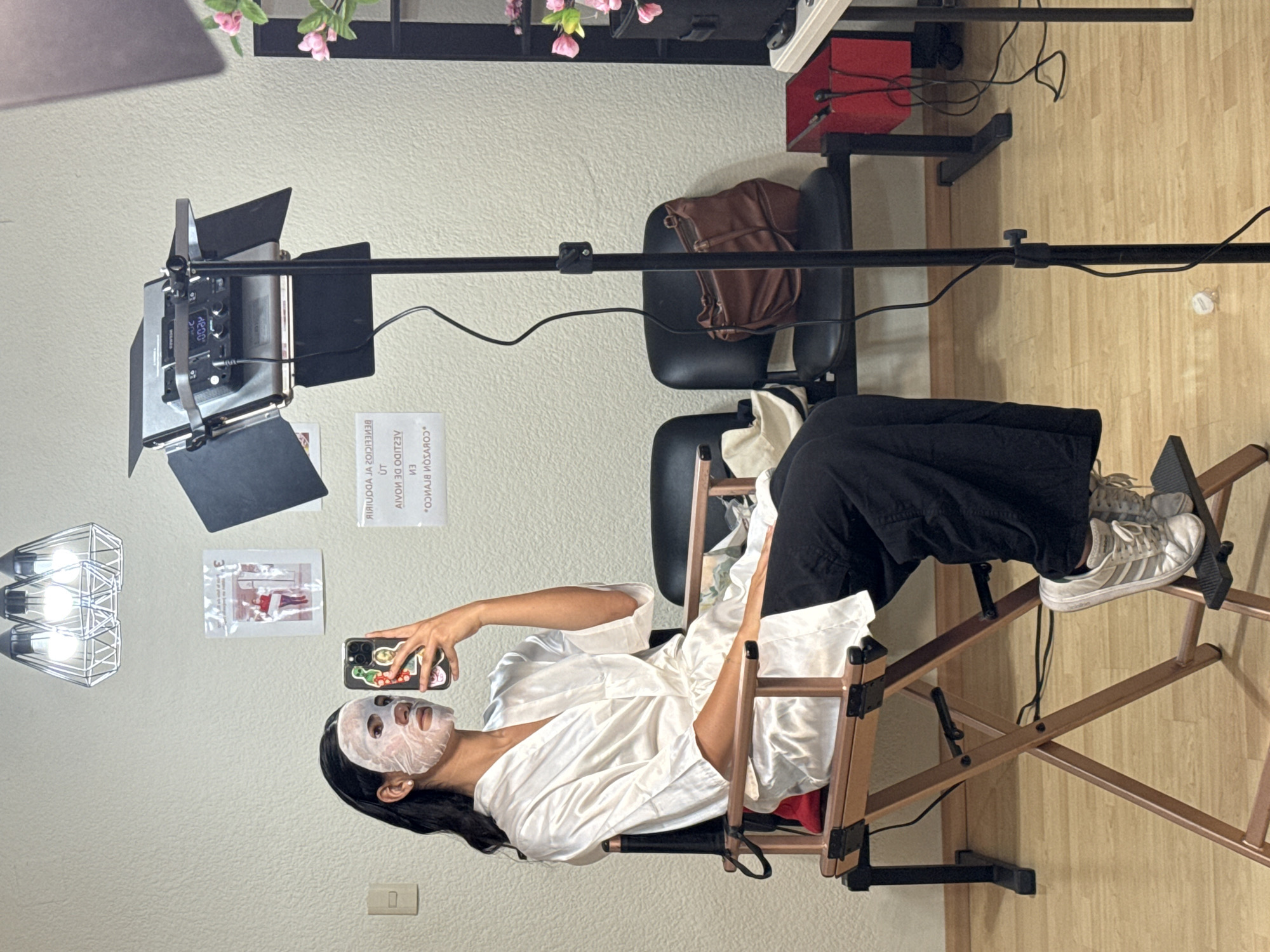 The image captures an indoor scene. A person with dark hair is seated in a director's chair wearing a white robe and a facial mask. They are holding a smartphone up as if taking a selfie. The chair is set on a wooden floor. Behind the chair are two black office chairs, one with a brown bag on it. Above the scene is a lighting rig with adjustable barn doors. On the wall behind the person is a light fixture and two advertisements. The image does not appear to contain information indicating that this photo was taken in Puebla, Mexico.