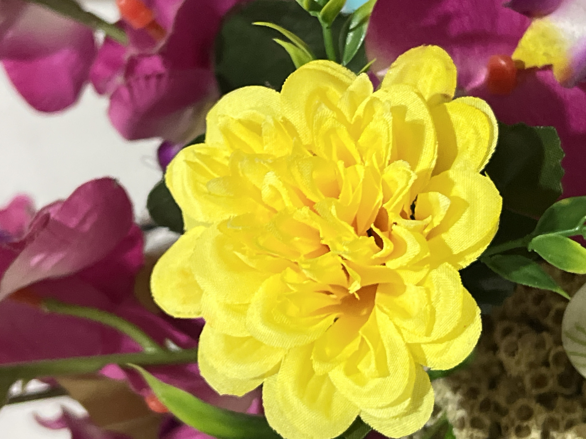 A close-up, eye-level shot focuses on a vibrant yellow, artificial dahlia flower. The flower's numerous, layered petals are tightly packed and textured, with a slightly ruffled appearance. The petals vary in shade from a bright yellow in the outer layers to a deeper, more golden hue towards the center, where the innermost petals are not fully visible. Surrounding the yellow flower are large, blurred, magenta-colored flower petals, which add a contrasting splash of color to the composition. Delicate green leaves and stems are also interspersed, with one pointed green leaf clearly visible in the lower left quadrant, extending towards the yellow bloom. The background is largely out of focus, with patches of white and hints of other colors, suggesting an artificial floral arrangement. The lighting is bright, highlighting the texture and color of the yellow flower. The image is devoid of any human presence or activity. The scene does not contain any specific information that allows for identification of its location within Denpasar, Indonesia.