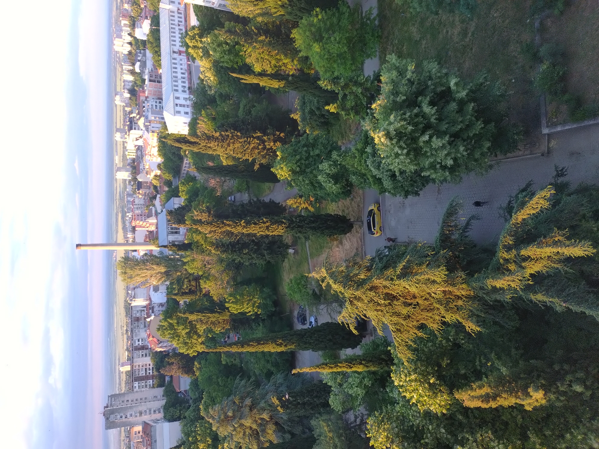 The image is a high-angle shot of an urban area in Stara Zagora, Bulgaria, heavily featuring lush greenery. A tall smokestack rises from the city landscape on the left, which transitions into the sky. Dominating the frame are numerous green trees of varying types, receiving ample sunlight. A paved road winds through the trees, hosting a parked yellow car and a lone pedestrian walking nearby. Further along the road, more parked cars are visible. Buildings are scattered in the backdrop, closer to the smokestack and towards the edge of the city.