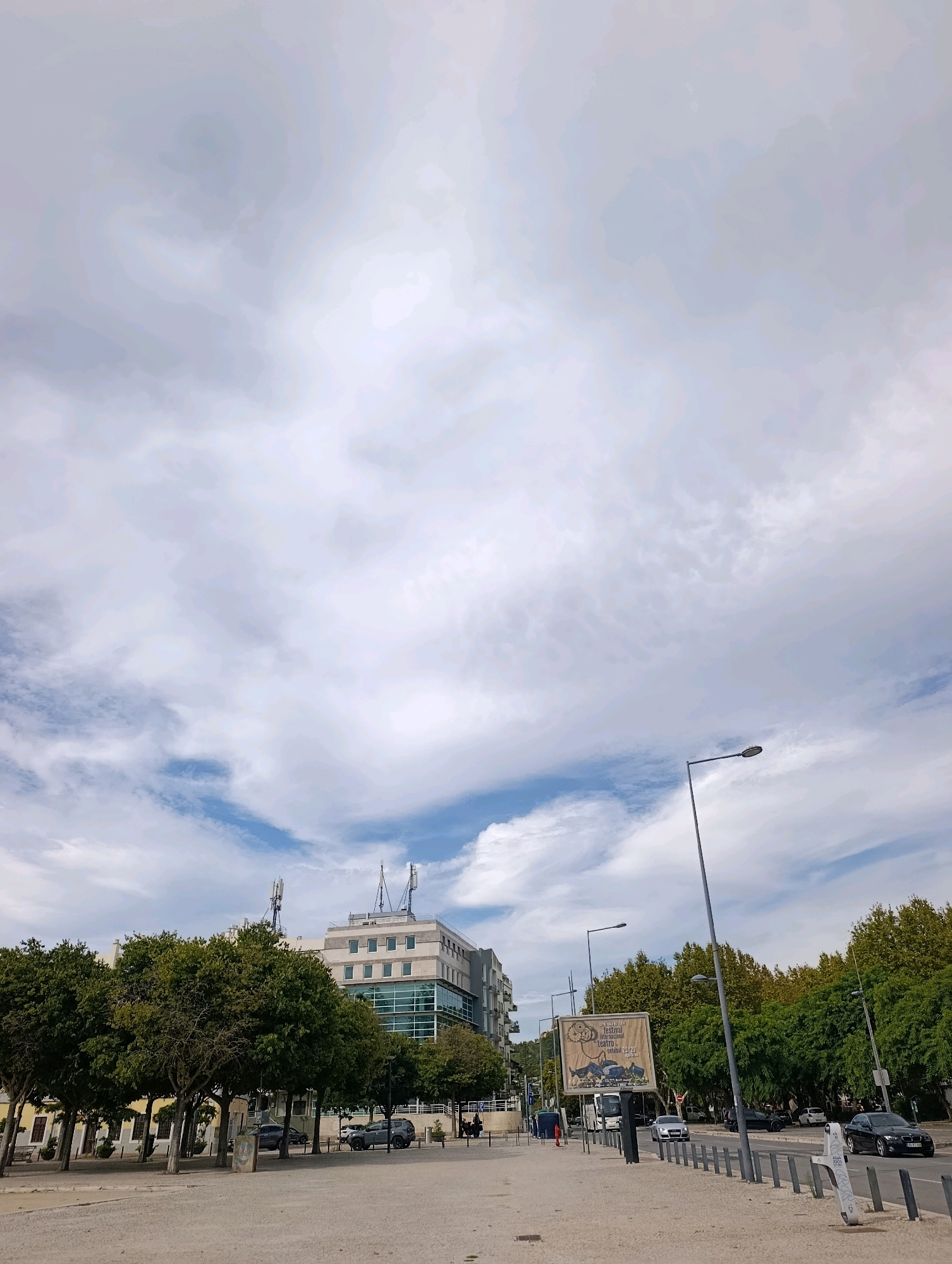 The image shows a street scene in Setúbal, Portugal, under a cloudy sky. In the foreground, a wide, paved area with scattered debris leads to a street lined with trees and buildings. A street lamp stands prominently on the right side of the image.
On the street, several cars are visible, including a black SUV and a silver sedan. A large billboard with an image of a building and text advertising a festival is positioned near the middle of the street. Buildings of various sizes and architectural styles line the street, some appearing to be residential or office buildings with multiple windows. Two cell towers are visible atop one of the larger buildings. The trees along the street are lush and green, providing shade.
The overall impression is a typical urban environment, with the cloudy sky suggesting a temperate climate. The scene appears to be captured during daylight hours. No people are clearly visible in the image.