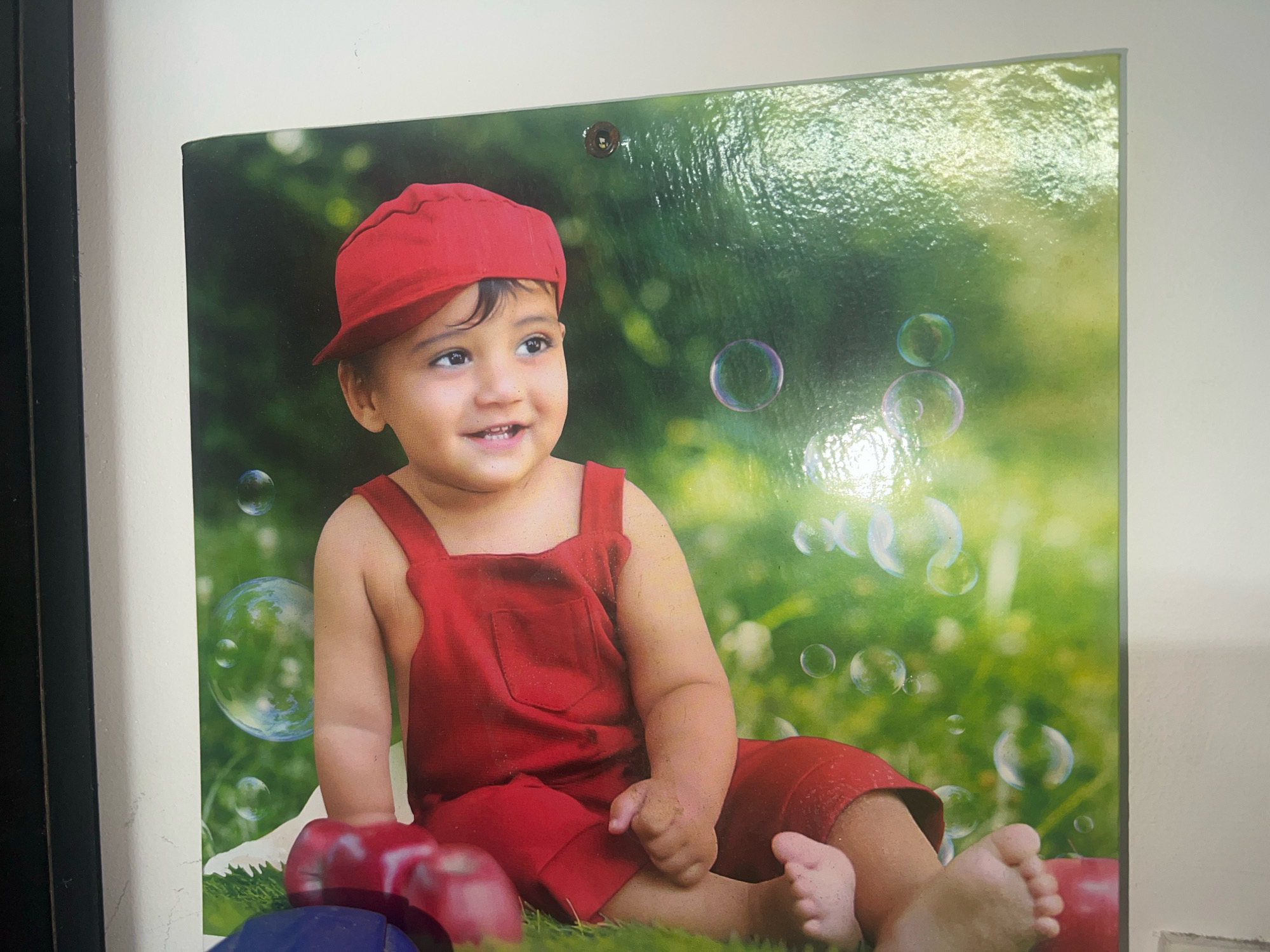 A framed photograph depicts a toddler sitting on a grassy surface, wearing a red cap and red overalls. The background is a blurred green landscape with several translucent bubbles scattered throughout, suggesting an outdoor or whimsical setting. The photograph is mounted on a light-colored wall, with a darker vertical element to its left. There are no discernible landmarks or context to indicate the location in Chennai, India.