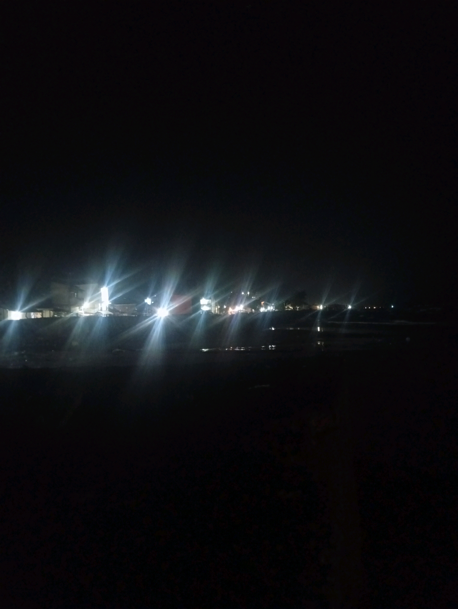 The image depicts a nighttime scene, likely a shoreline or a body of water, with a faint outline of buildings and lights visible in the distance. The sky is dark, indicating it is night. The lights from the buildings are reflected on the water's surface, creating a shimmering effect. The foreground is dark and indistinct, suggesting it might be a beach or a pier. The overall impression is one of a quiet, perhaps tranquil, coastal area at night. No people or specific landmarks are identifiable in the image.