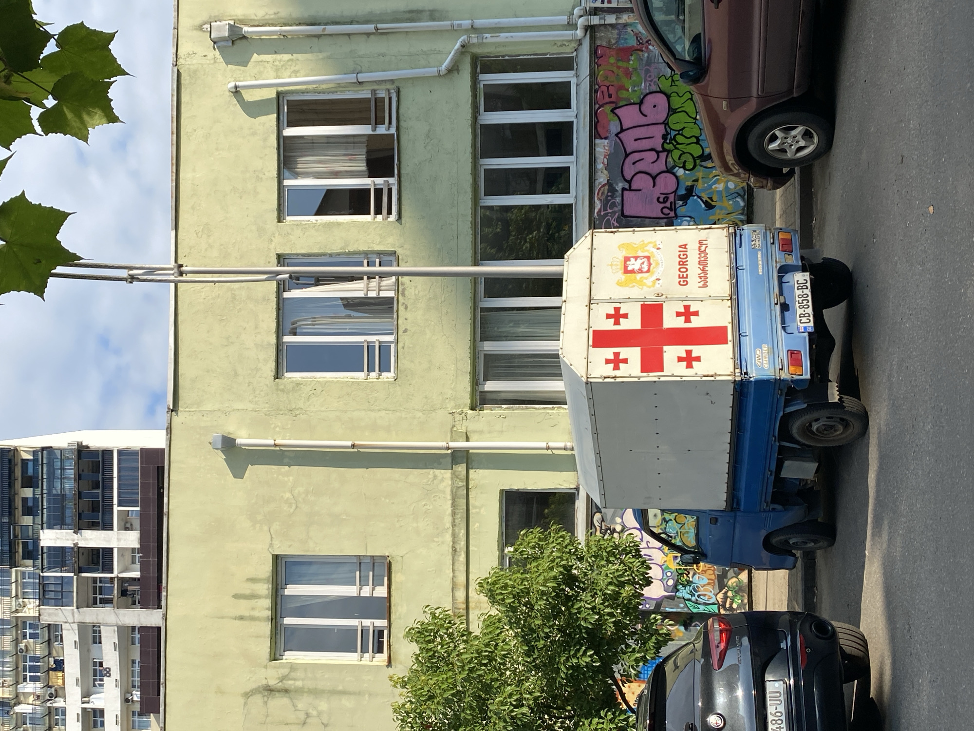 The image depicts a street scene in Batumi, Georgia. In the foreground, a blue and white utility truck is parked, featuring the flag of Georgia and the word "GEORGIA" in English and Georgian script on its rear cargo box. To the left of the truck, the rear of a black SUV is visible. To the right, a brown car is parked. The background consists of a pale green building with multiple windows, showing graffiti on its lower facade. Above the green building, a tall, white residential building with many balconies and windows dominates the upper part of the frame. Lush green foliage from a tree is visible on the left side of the image, and the sky is partly cloudy.