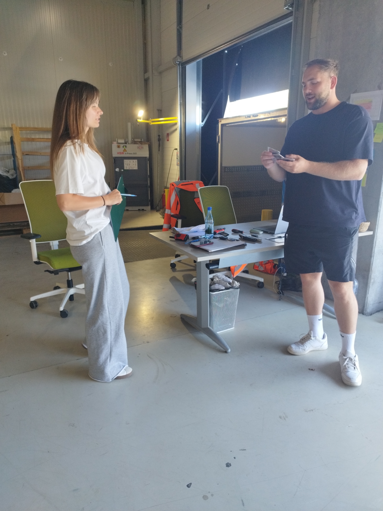 In Cernica, Romania, two individuals, a woman and a man, stand near a desk in what appears to be a warehouse or industrial setting. The woman, facing left, wears a white t-shirt and grey sweatpants, holding a green folder. The man, facing the woman, is dressed in a navy t-shirt and black shorts, holding a small object in his hands. A desk between them is cluttered with various items including a laptop, bottles, tools, and other office supplies. Two green chairs are positioned near the desk, and a trash can is visible. The background features industrial elements such as metallic walls, overhead lighting, and large doors.