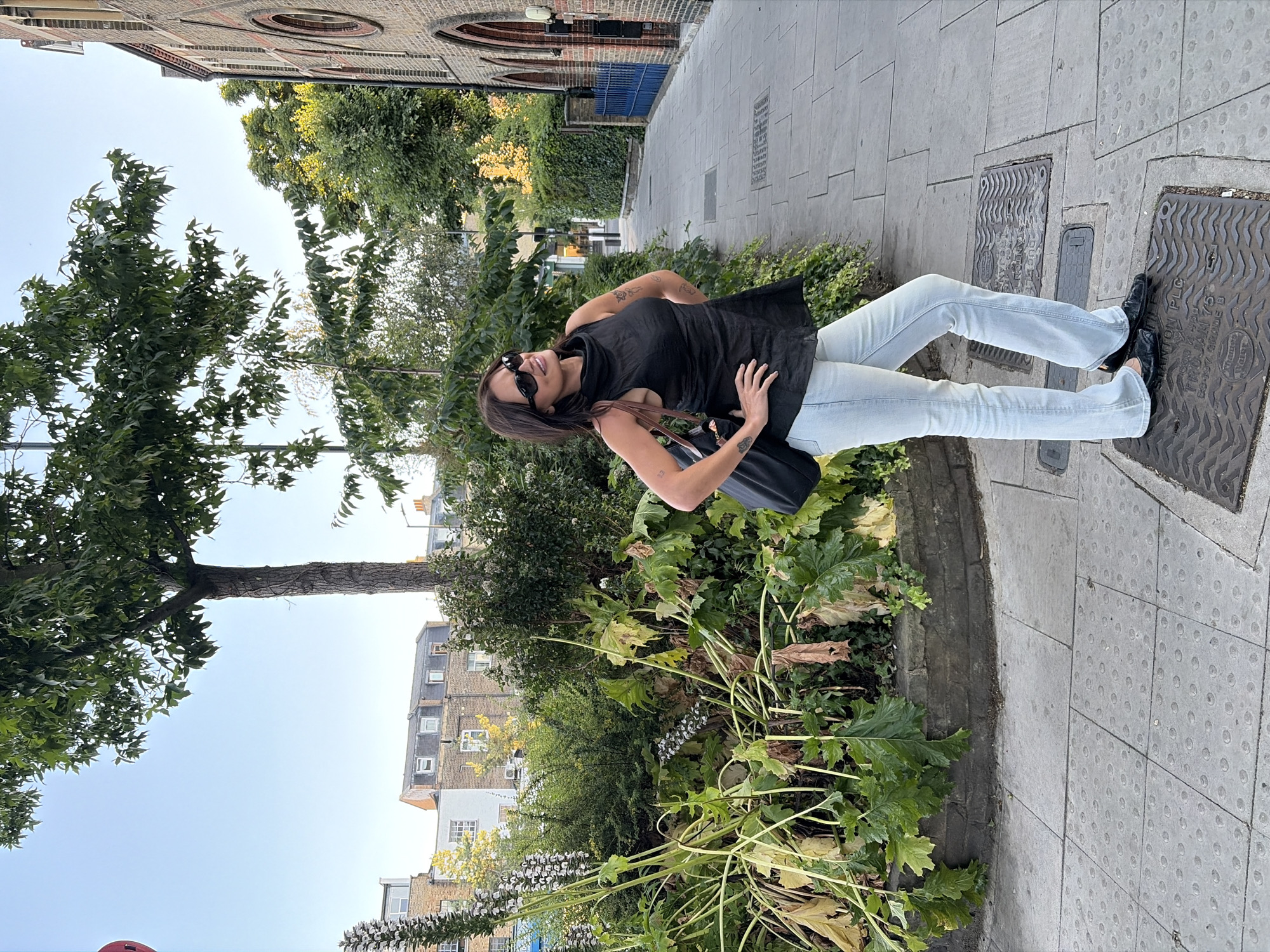 A woman stands on a paved sidewalk next to a garden bed filled with large-leafed plants and flowers. She is wearing a black sleeveless top, light blue flared jeans, black shoes, and sunglasses. She has her left hand on her hip and her right arm is bent. A black handbag is slung over her right shoulder. Tattoos are visible on her arms. Behind her, a narrow paved pathway leads to a brick building with arched windows, and trees and greenery line the path. The sky is clear and blue, suggesting daytime. The presence of textured paving slabs on the sidewalk indicates a public outdoor space. Buildings with brick facades and multiple windows are visible in the background, consistent with an urban environment like London. The overall mood appears casual and relaxed.