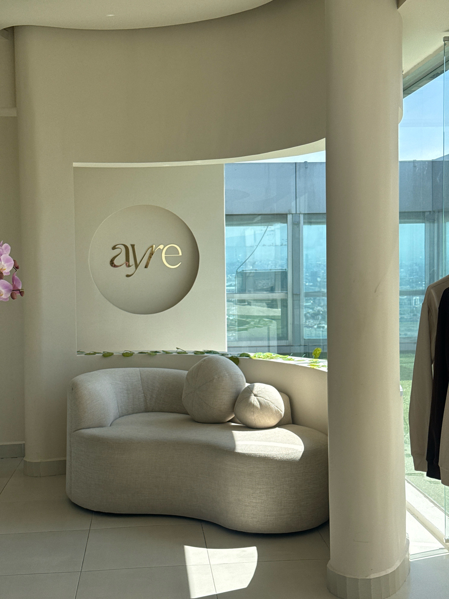 The image depicts an interior scene with a curved, light-colored sofa positioned in front of a wall. A circular emblem with gold lettering that reads "ayre" is mounted on the wall above the sofa. To the left of the sofa, a branch with pink orchids is visible. The room features large windows offering a view of a cityscape, suggesting an urban location. A thick, cream-colored pillar stands to the right of the sofa, adjacent to the windows. A garment, possibly a jacket, is partially visible hanging on the pillar. The floor is tiled in a neutral tone. The lighting suggests it is daytime, with sunlight casting shadows on the floor. The presence of the "ayre" emblem indicates this may be a commercial or branded space. The setting appears to be a modern, minimalist interior.