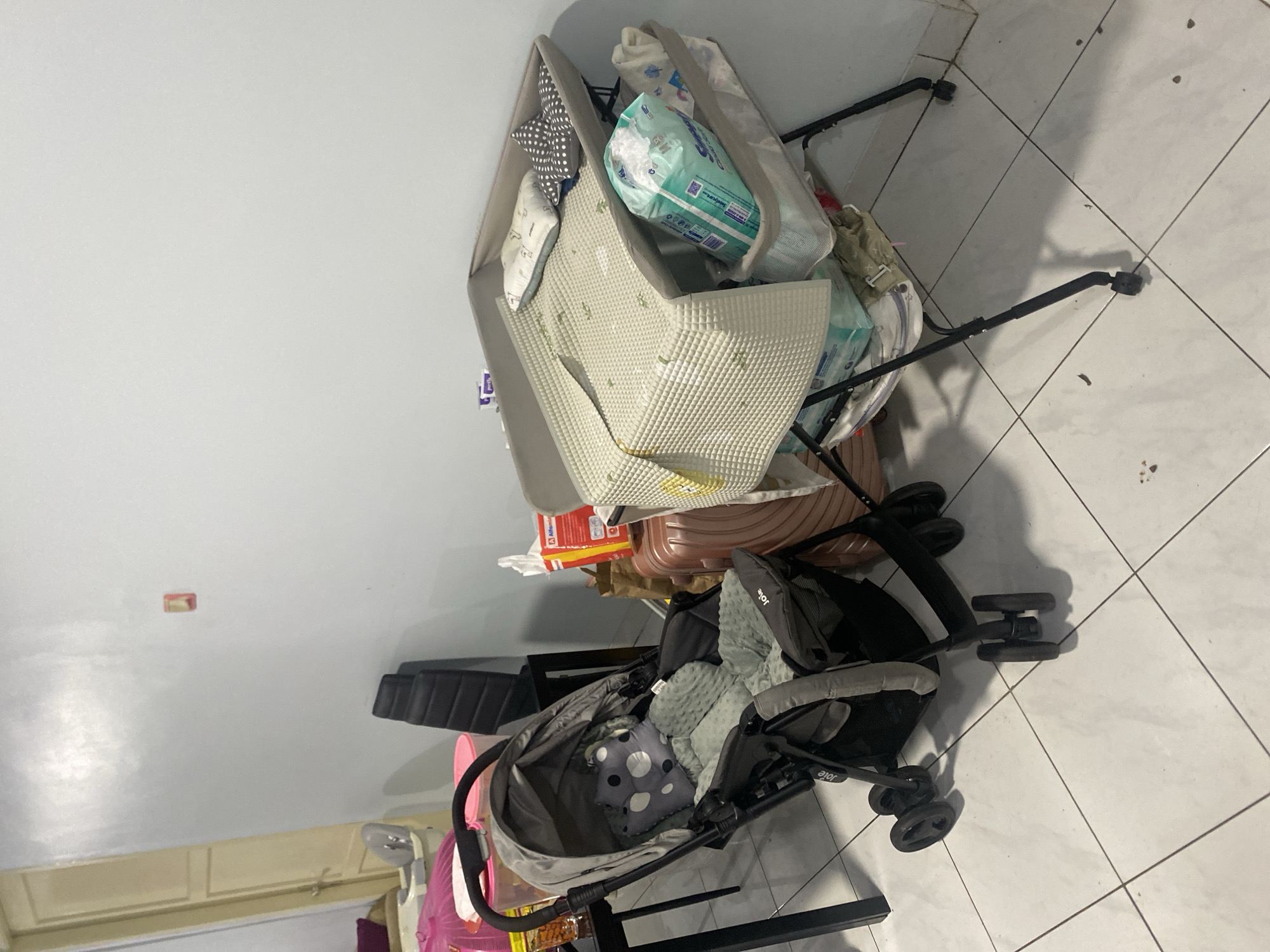 The image depicts an indoor scene, likely a room in a residential setting, potentially in Dukuh, Indonesia, given the context. The primary focus is on a baby changing station and a stroller. The changing station is gray with black legs and has a padded surface covered by a light-colored blanket with a subtle green pattern. A small white pillow and a gray pillow with white polka dots are placed on the changing surface. Below the changing surface, there is a storage compartment containing a large package of diapers, a white towel, and other unidentifiable items.

Adjacent to the changing station is a gray stroller, also with black accents. The stroller is equipped with a canopy and has a padded seat with a green patterned mat and a gray cushion with white polka dots. The brand "Joie" is visible on the stroller.

In the background, a pink object, possibly a baby bath, is partially visible on a table. To the right of the changing station, a large, brown suitcase is present. A black office chair is also visible in the mid-background, leaning against the wall. The walls are painted a light color, and the floor is tiled with white squares. The lighting suggests an indoor environment, possibly with artificial light. No people are visible in the image. The overall impression is of a space organized for childcare, with essential baby items readily available.