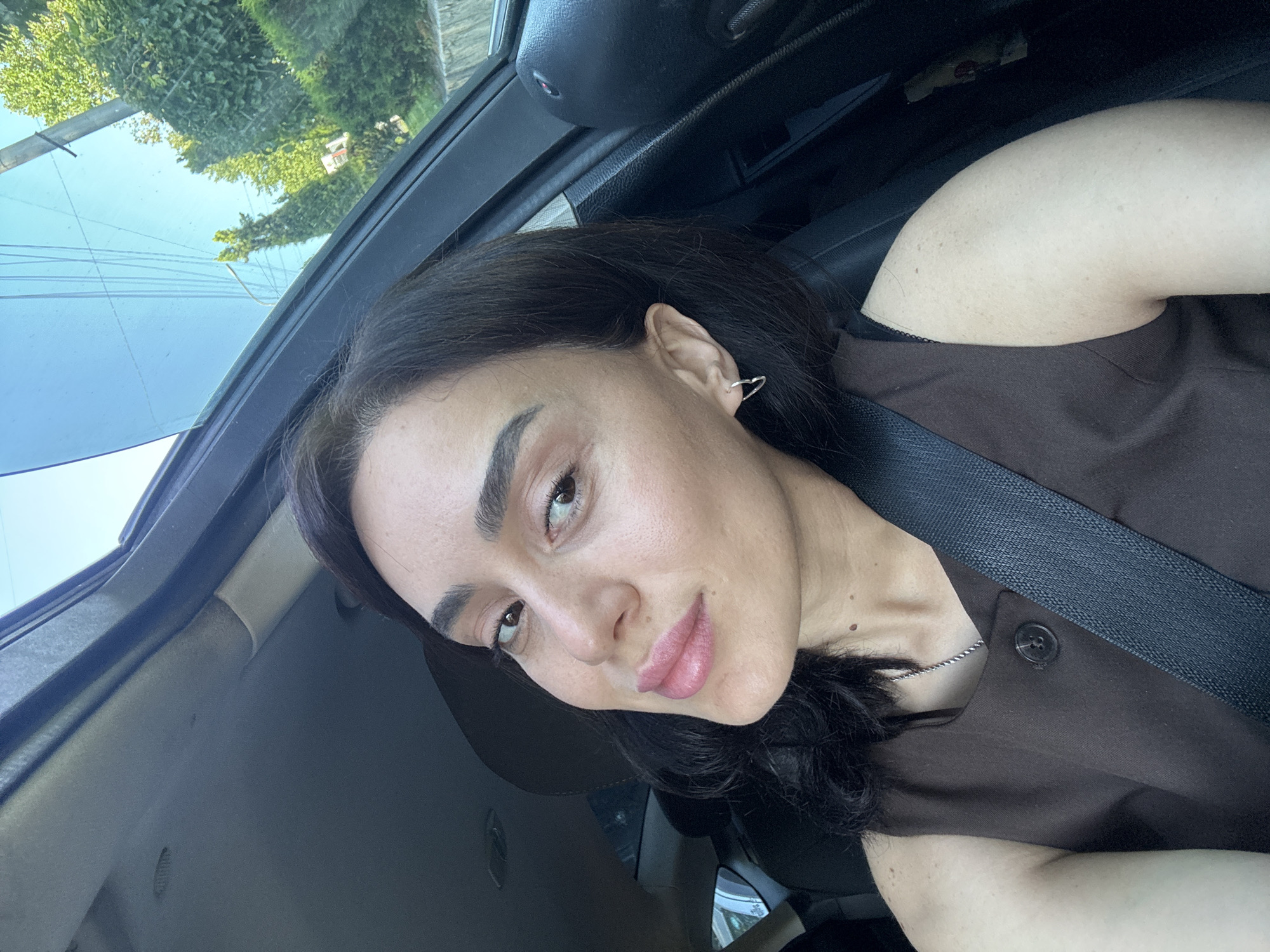 A woman with dark hair and makeup is taking a selfie in a car. She is wearing a brown sleeveless top and a seatbelt. The car interior is visible, as is the sky and trees through the open window. The location is in Tbilisi, Georgia.