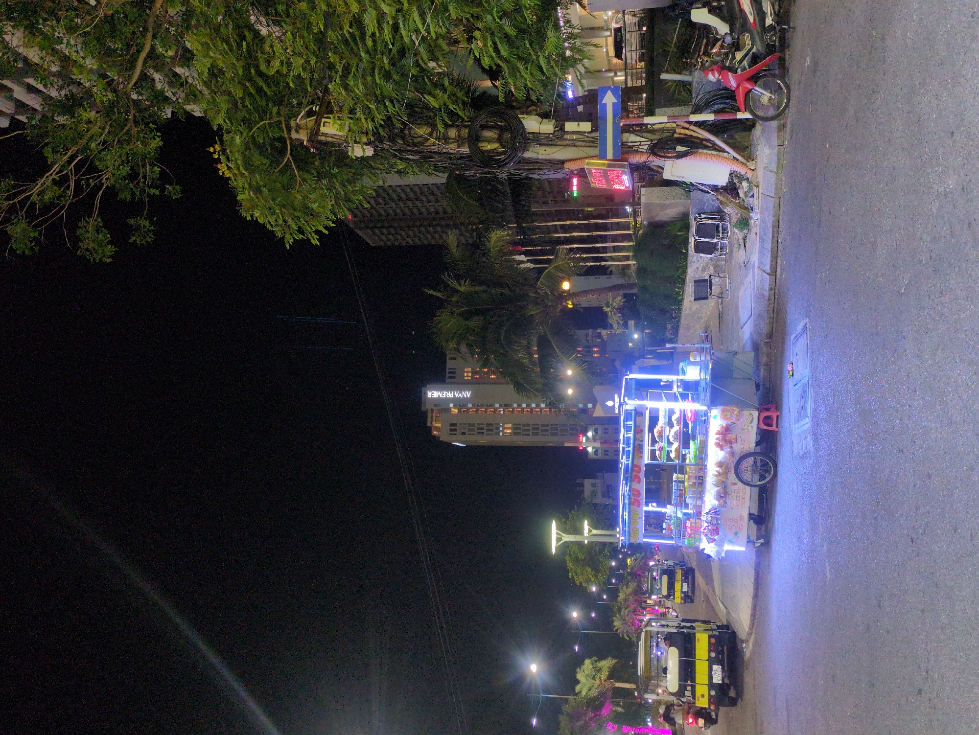 A nighttime scene in South Quy Nhon Ward, Vietnam, captures a vibrant urban atmosphere. The foreground shows a paved street with several vehicles. A brightly illuminated food cart stands out with its blue and white lighting. Nearby, auto rickshaws are parked along the curb, some with visible license plates. The backdrop reveals buildings of varying heights, including the 'Anya Premier' building, illuminated by numerous lights. The surroundings include trees and street lighting, adding to the urban landscape. Overhead, there is a complex network of utility lines and equipment. A parked red scooter is visible. The overall scene conveys a bustling nightlife in the city.