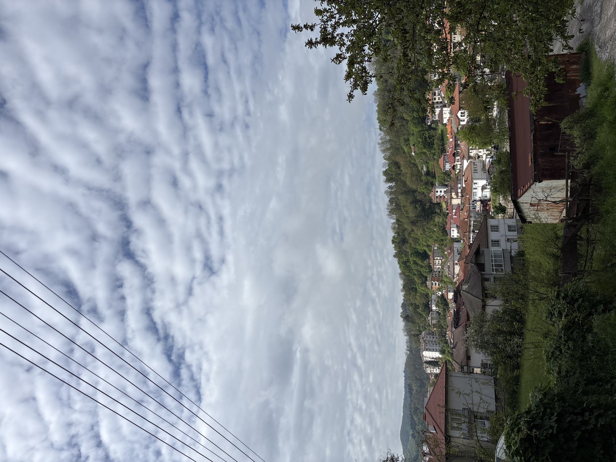 The image depicts a street scene in what appears to be Zlatograd, Bulgaria, characterized by houses situated on a hillside. The sky is filled with a pattern of cirrocumulus clouds. Power lines are visible in the lower left corner. The hillside is densely populated with houses, showing a variety of architectural styles and red-tiled roofs. Some green foliage and trees are interspersed among the houses. A rusty structure is visible on the right side of the image.