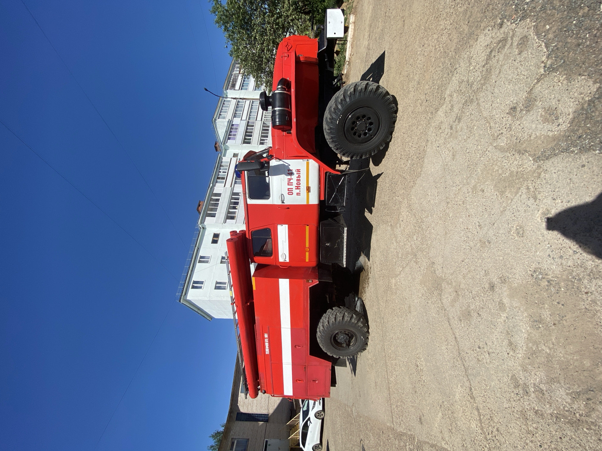 A red and white fire truck with Russian text on the side reading "ОП ПЧ-41 п. Новый" is parked on a concrete surface. The building behind the fire truck appears to be an apartment complex. The sky is blue. A white car is parked partially out of frame to the right.
