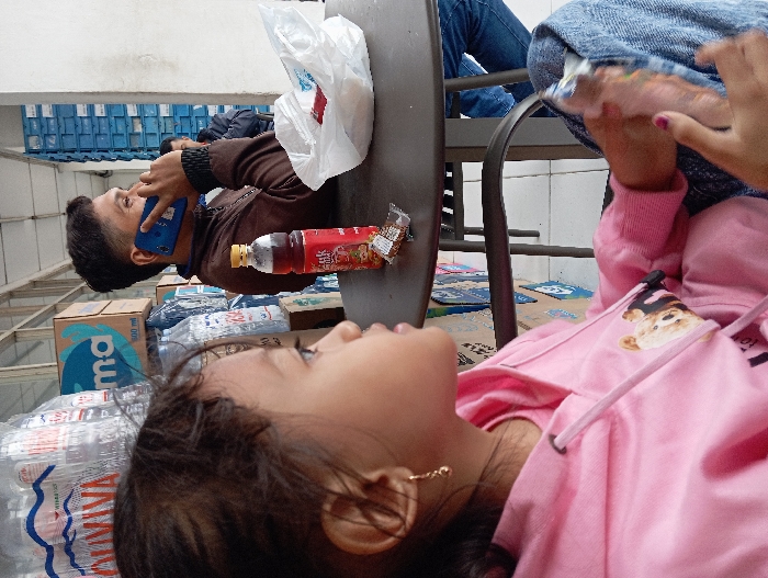 The image depicts an outdoor seating area in Legok, Indonesia, featuring three individuals seated at a dark-colored, round, slatted table. In the foreground on the left, a young female child, estimated to be 4-7 years old, is seen in profile, facing right. She wears a pink hooded sweatshirt with a graphic depicting two bears and blue denim jeans. A small gold-colored hoop earring is visible in her left ear. Her right hand rests on her lap, holding a clear plastic-wrapped snack.

In the midground, to the child's right, an adult male is seated at the table, also in profile facing right. He wears a brown long-sleeved jacket and is engaged in a phone call, holding a blue smartphone to his right ear. On the table in front of him are a red plastic beverage bottle with a yellow cap, a small brown foil-wrapped item, and a white plastic bag containing a red object.

A third adult male is partially visible in the background, seated behind the first male. The background features large stacks of bottled water containers, some labeled "VIVIVA" and "QUAVIVA," alongside cardboard boxes, one marked with "ma" and "800 ml." To the far right, a vertical stack of blue plastic crates is visible against a light-colored wall.