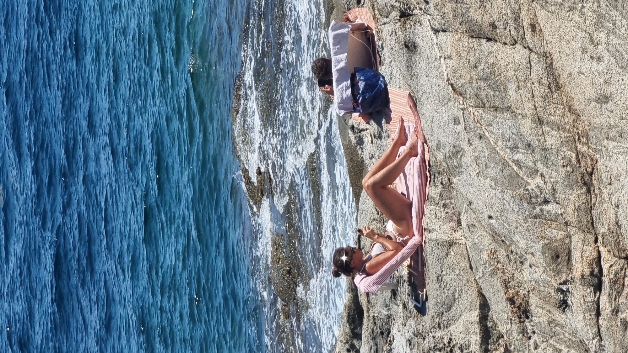 Two individuals are sunbathing on a rocky shoreline next to the Mediterranean Sea in Roses, Spain. The person in the foreground, wearing a white bikini, is reclining on a pink striped beach towel and a folded pink cushion. They are holding and looking at a smartphone. Their legs are extended towards the viewer. In the background, a second person is lying on their side on a white towel, facing away from the camera, with their head resting on a white pillow. A blue bag is placed next to them. The sea is a vibrant blue with white waves breaking against the rocky shore. The terrain is characterized by rough, grey rocks. The sun is shining brightly, casting shadows on the rocks.