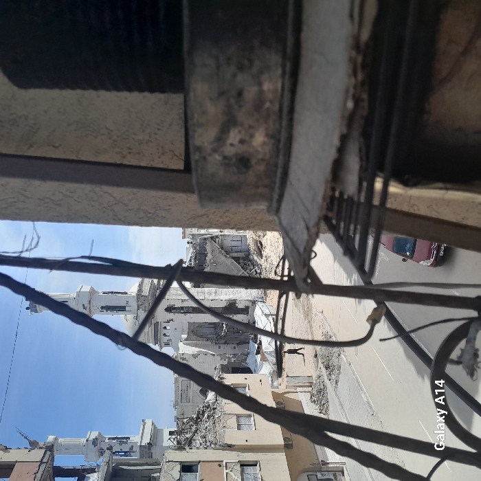 The image depicts a street scene in Khan Yunis, Palestinian Territory, viewed from an elevated perspective, possibly a window or balcony, partially obstructed by a dark, weathered metallic structure in the immediate foreground on the right.
The central feature is a severely damaged multi-domed mosque with two prominent minarets, its walls heavily pockmarked, with large sections of its structure, including the roof and interior, exposed or collapsed into rubble. Surrounding the mosque are numerous other buildings, also extensively damaged or reduced to piles of debris. The sky above is clear and blue.
A paved road runs through the midground. On the left side of the street, a lone individual, dressed in dark attire, walks away from the viewer towards the damaged mosque. On the right side of the street, a red compact car, identifiable by its silhouette and a visible portion of its license plate, moves towards the viewer. Rubble and construction debris are scattered along the road edges and around the damaged structures. The timestamp or watermark "Galaxy A14" is visible in the bottom left corner.