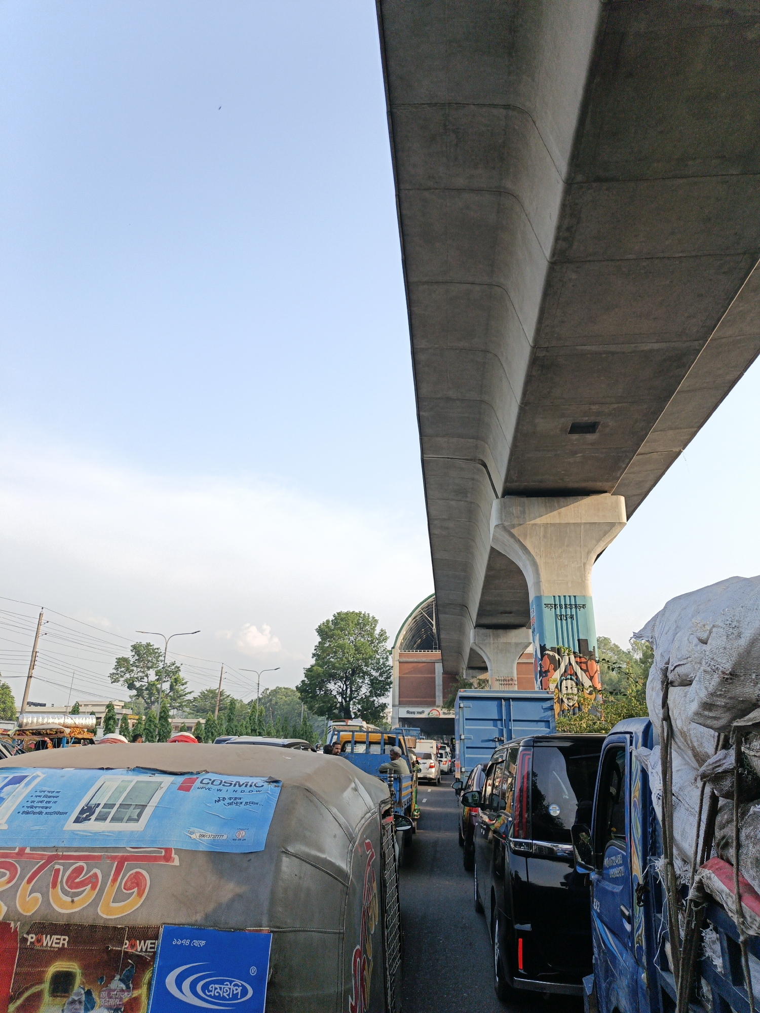 The image shows a congested street scene in an urban environment, likely in Asia based on the vehicles and writing. A large concrete elevated structure, possibly a metro line or highway overpass, dominates the upper right of the frame. The support columns for the structure are painted with colorful artwork. Below the structure, dense traffic is visible, including motorized rickshaws ("easy bikes") and cars. The vehicles are packed closely together, suggesting a traffic jam. The sky is light blue with some clouds, and trees and streetlights are visible in the background. Signage is present, with some words in Bengali. The OCR reveals text like "COSMIC UPVC WINDOW", "POWER", "বিজয় সরণি" (Bijoy Sarani, Victory Avenue), and "সড়ক ও মহাসডক" (Roads and Highways Department).